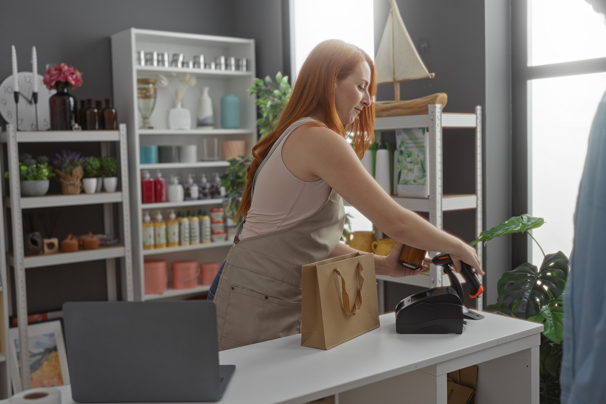 Lojista ruiva de avental passando um item no leitor de código de barras, no balcão de uma loja de decoração com prateleiras de vasos, velas e plantas ao fundo.