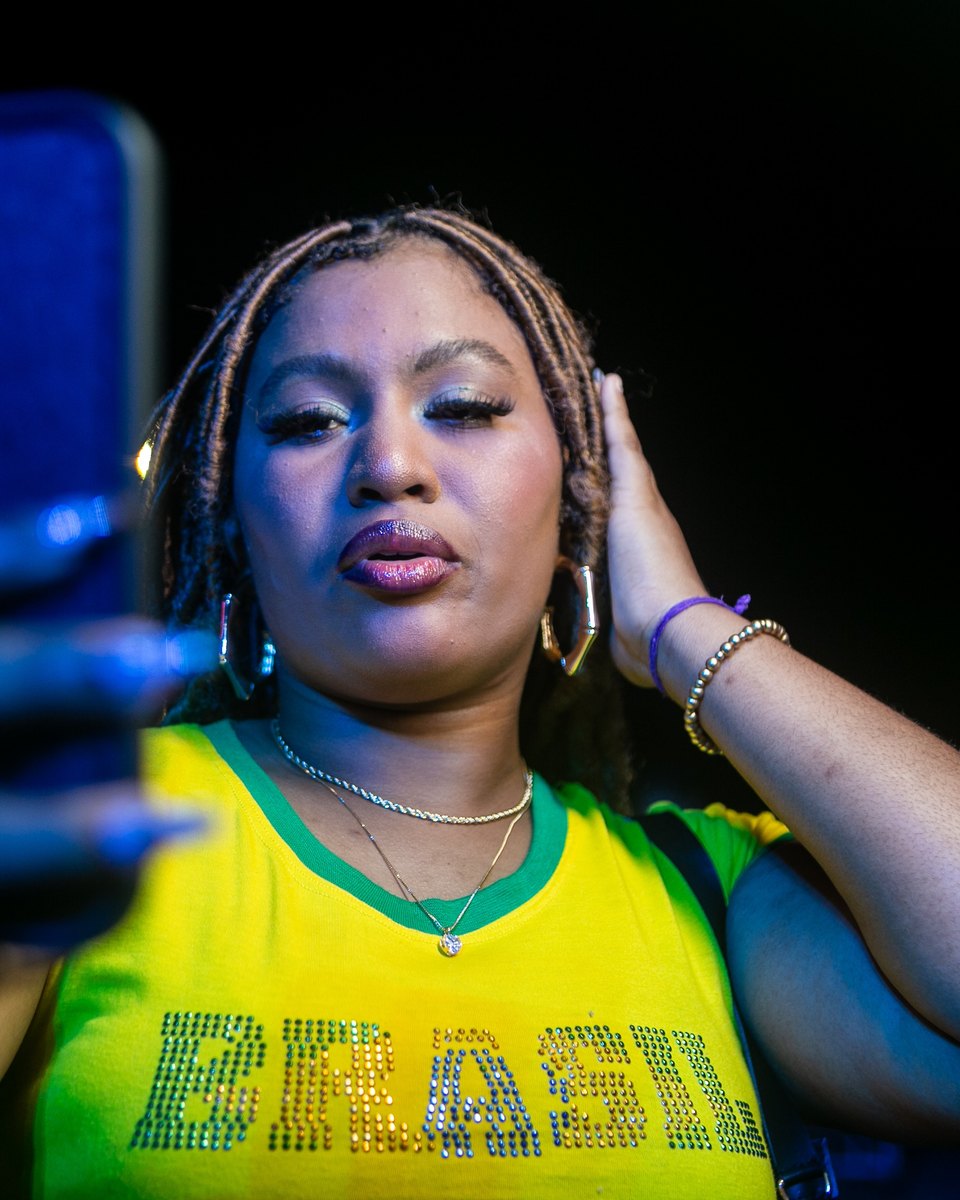 Jovem negra de tranças, argolas douradas e blusa amarela e verde escrita Brasil fazendo uma selfie com o celular à noite, no AFROPUNK, no Parque de Exposições de Salvador.