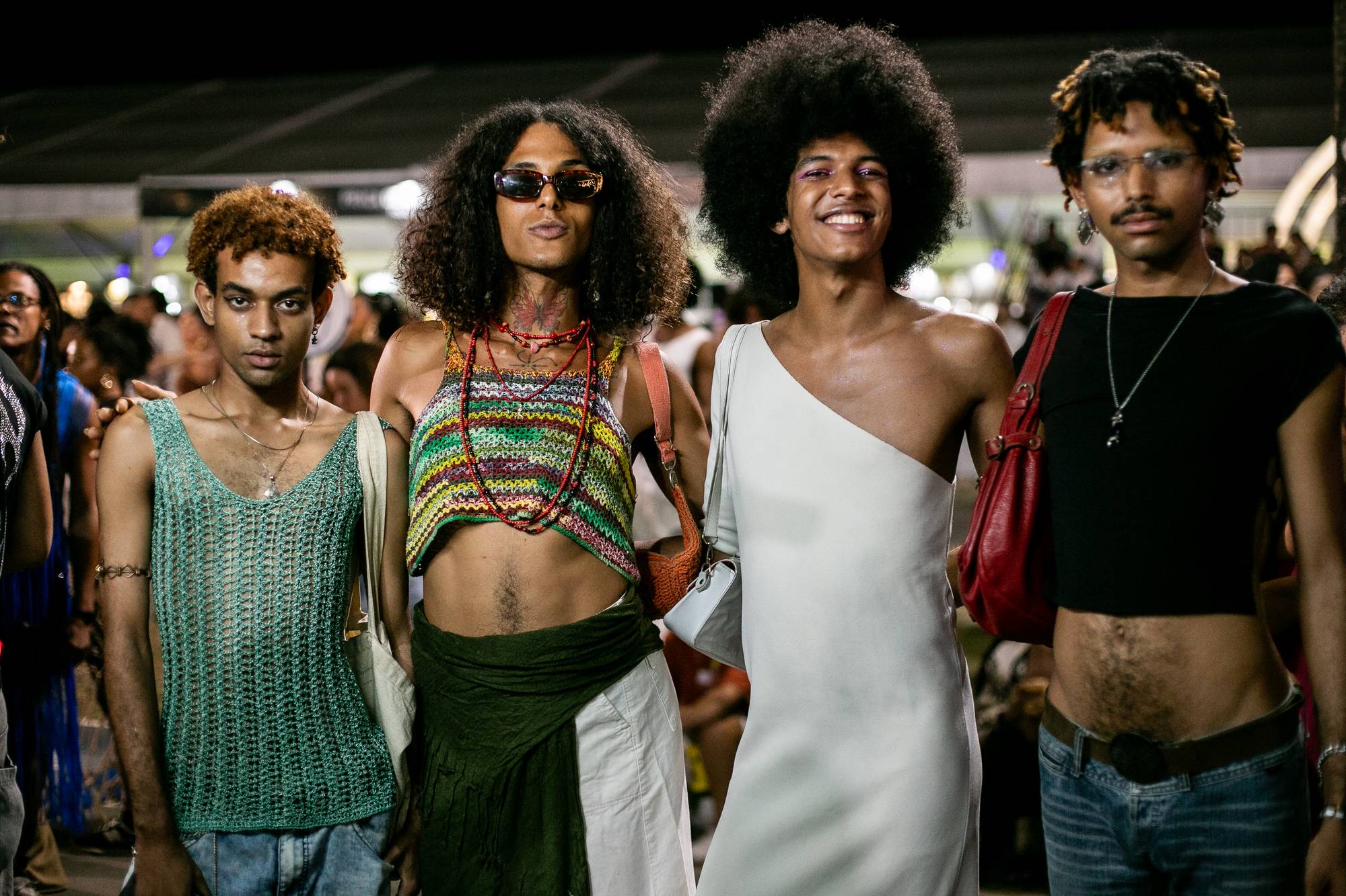 Quatro jovens negros posando lado a lado à noite no AFROPUNK, no Parque de Exposições de Salvador, com cabelos black power e cacheados, blusa de crochê colorida, vestido branco de um ombro só, colares e óculos escuros, e o público do festival ao fundo.