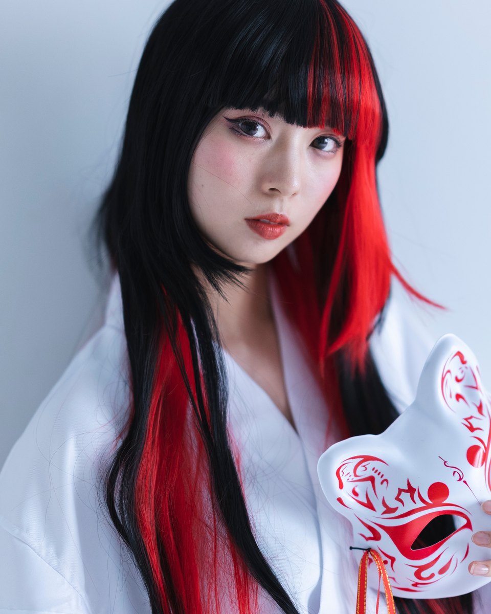 Jovem de peruca preta com mechas vermelhas e traje branco de sacerdotisa japonesa segurando uma máscara de raposa pintada de vermelho.