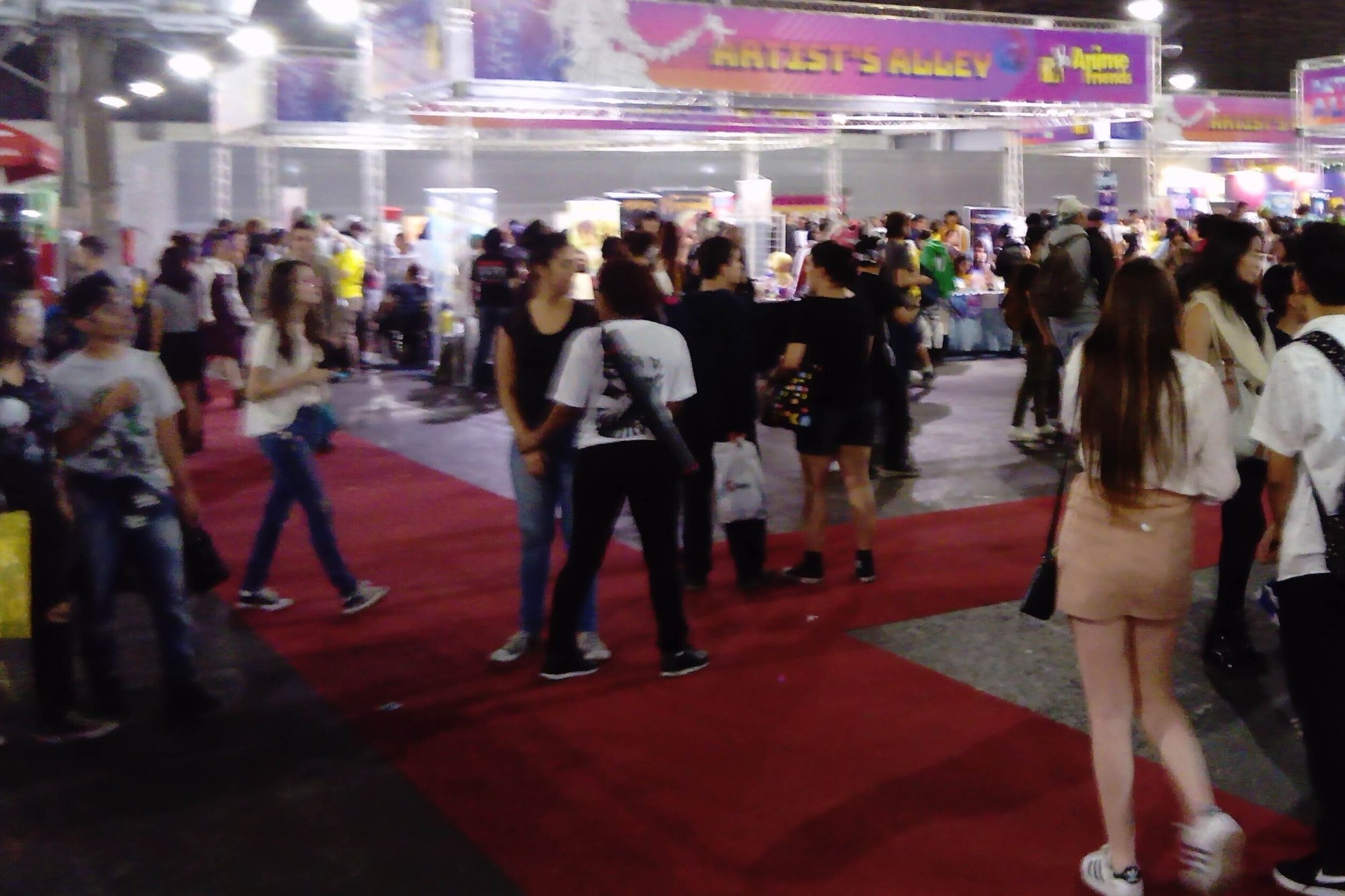 Jovens de mochila e sacola circulando no tapete vermelho do pavilhão do Anime Friends de 2018, em São Paulo, com o corredor de artistas independentes ao fundo.