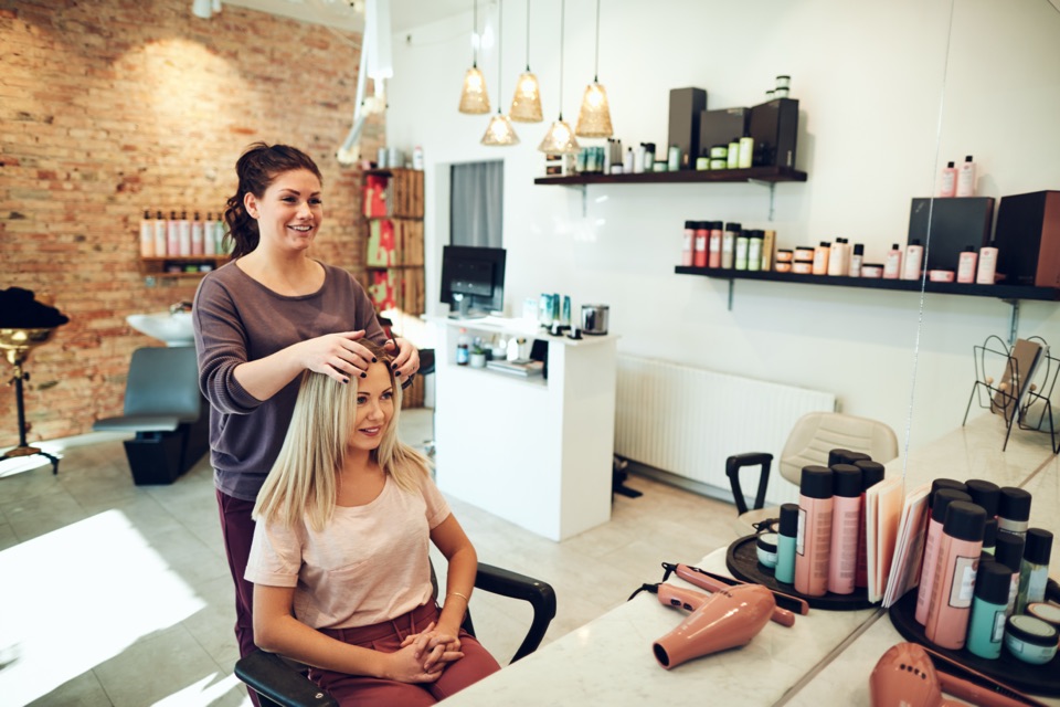 Cliente sorrindo e conversando com a cabeleireira num salão de beleza claro, com prateleiras de produtos ao fundo.