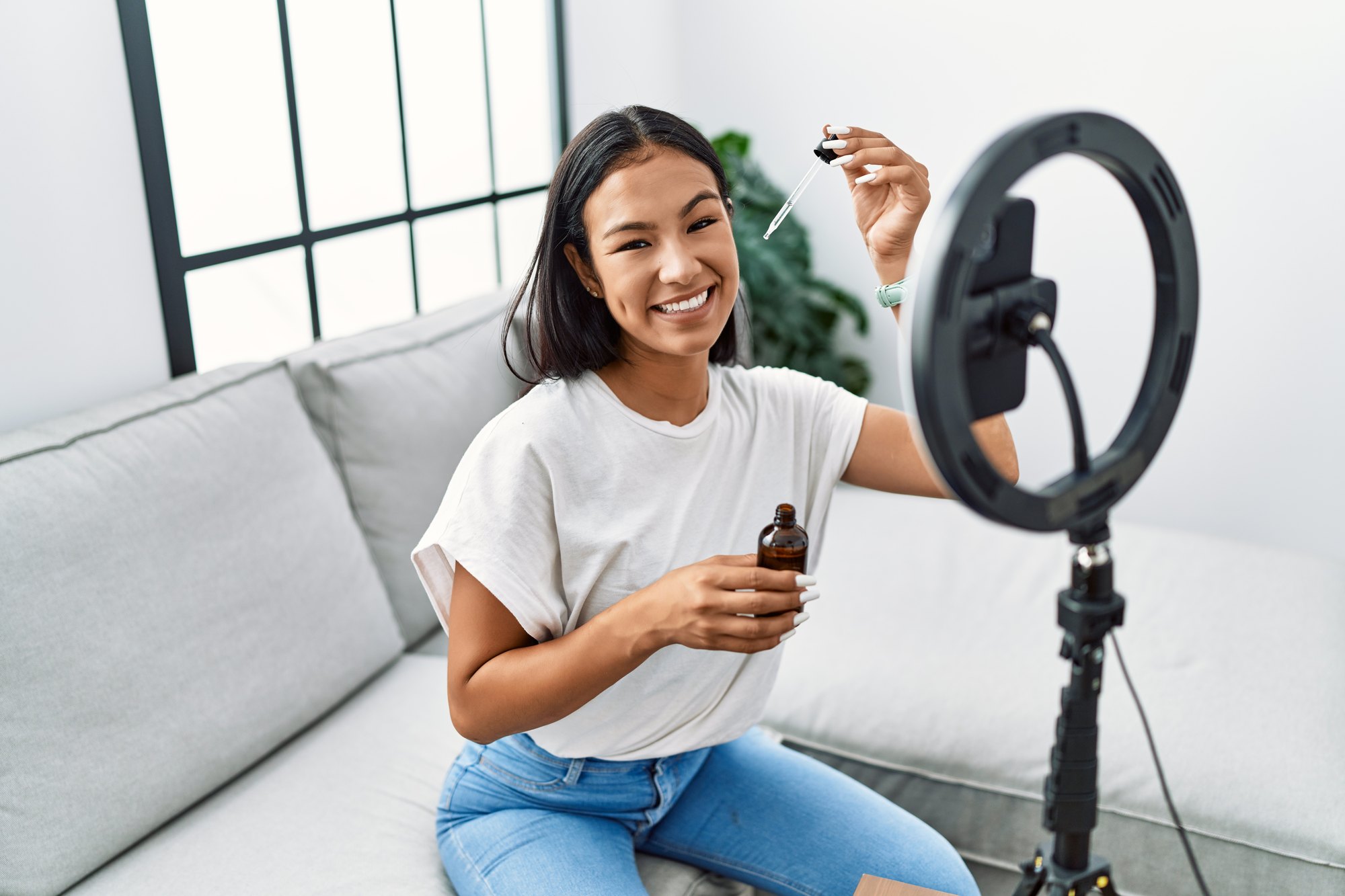 Jovem de cabelo escuro sorrindo e segurando um frasco de sérum com conta-gotas diante de um celular preso num ring light, gravando um vídeo na sala de casa.