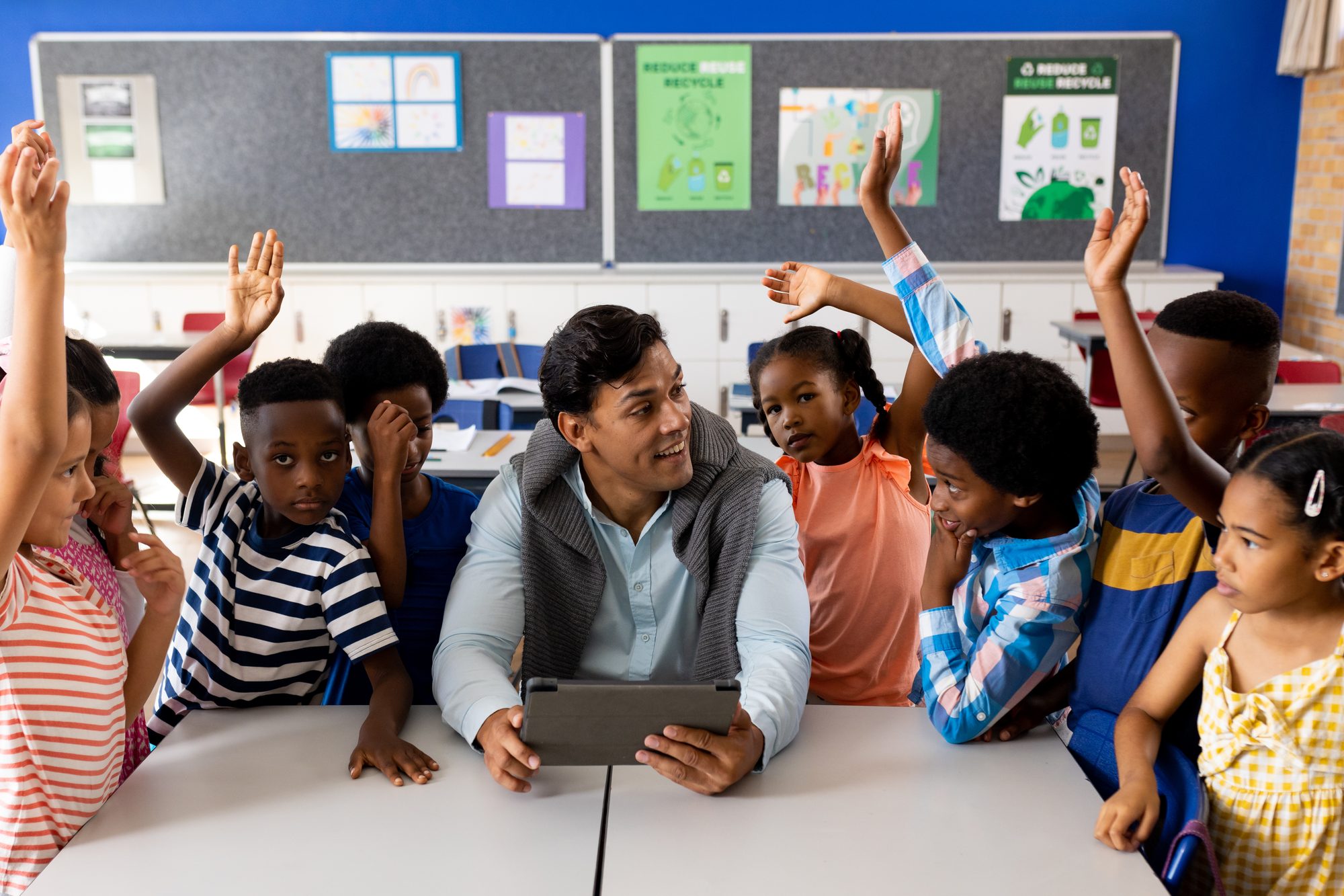 Professor sentado numa sala de aula com um tablet nas mãos, cercado de alunos do ensino fundamental que levantam a mão para responder.