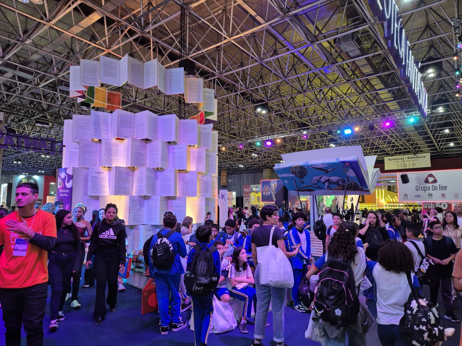 Pavilhão lotado da Bienal do Livro do Rio de Janeiro em 2025, com uma torre de páginas de livro abertas e iluminadas e estudantes de uniforme azul circulando.