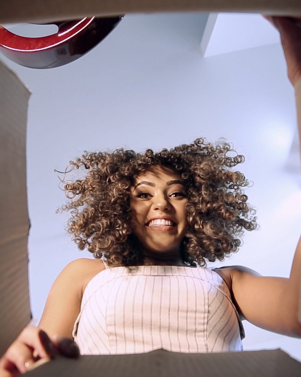 Mulher de cabelo cacheado sorrindo ao abrir uma caixa de encomenda, vista de dentro da caixa.