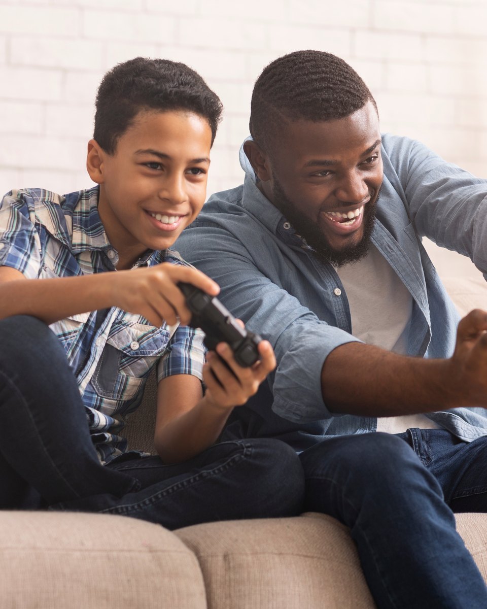 Pai e filho sentados no sofá de casa, rindo e disputando uma partida de videogame, cada um com o seu controle na mão.