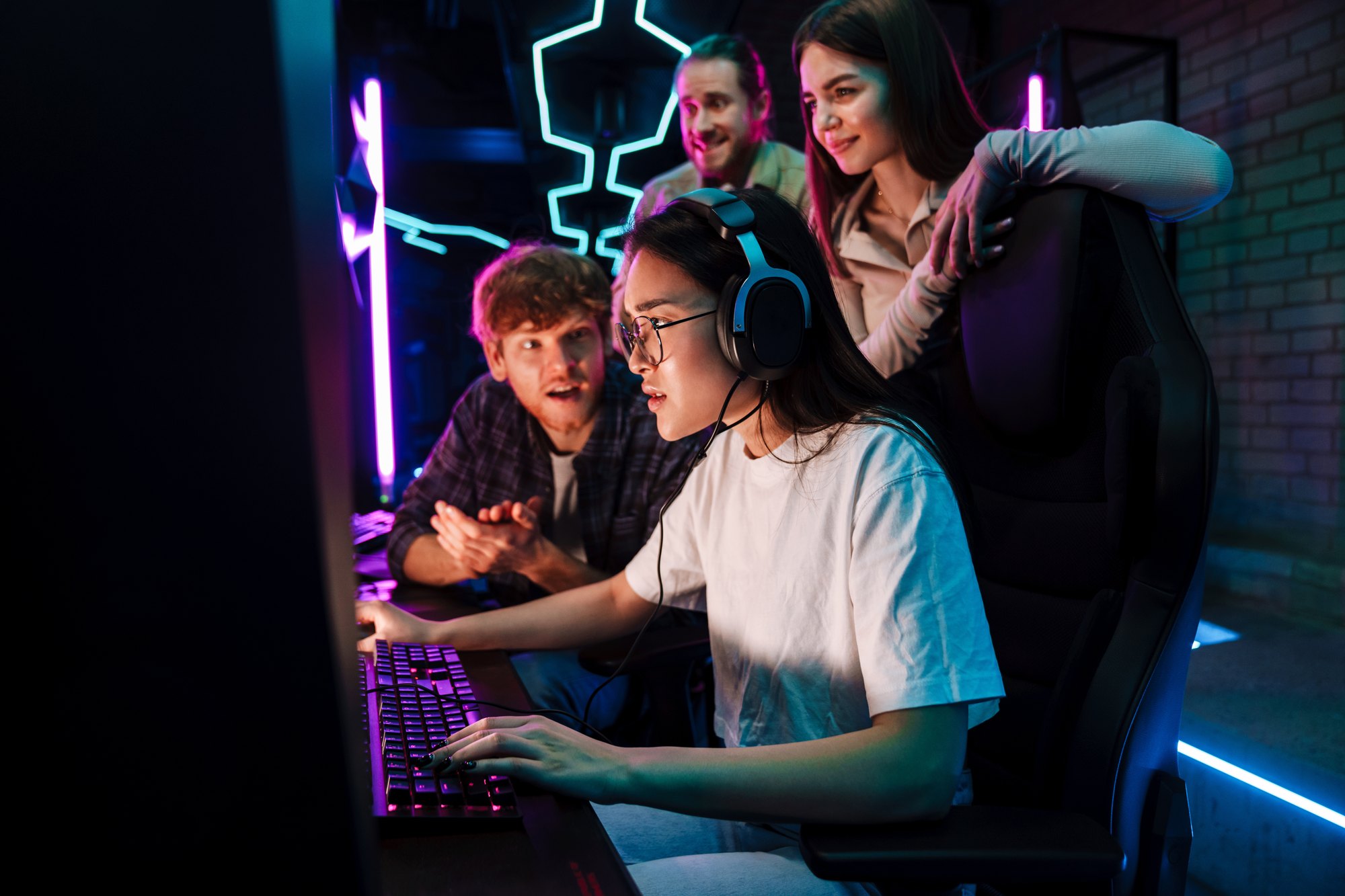 Jovem de fone de ouvido jogando no computador numa sala iluminada de neon roxo e azul, com três amigos em volta torcendo e dando palpite.