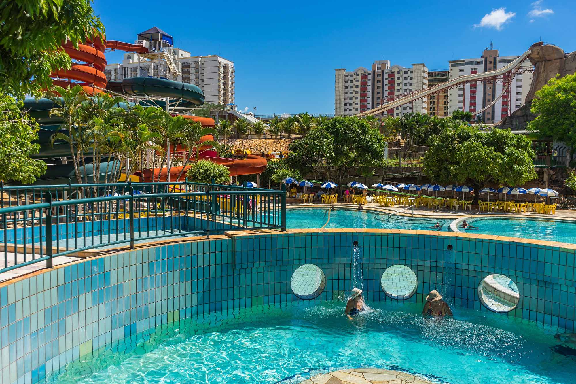 Duas pessoas de chapéu de sol sentadas sob as bicas de uma piscina termal de azulejos azuis no diRoma Acqua Park, em Caldas Novas, com guarda-sóis, toboáguas laranja e prédios de hotel ao fundo, num dia de céu azul.