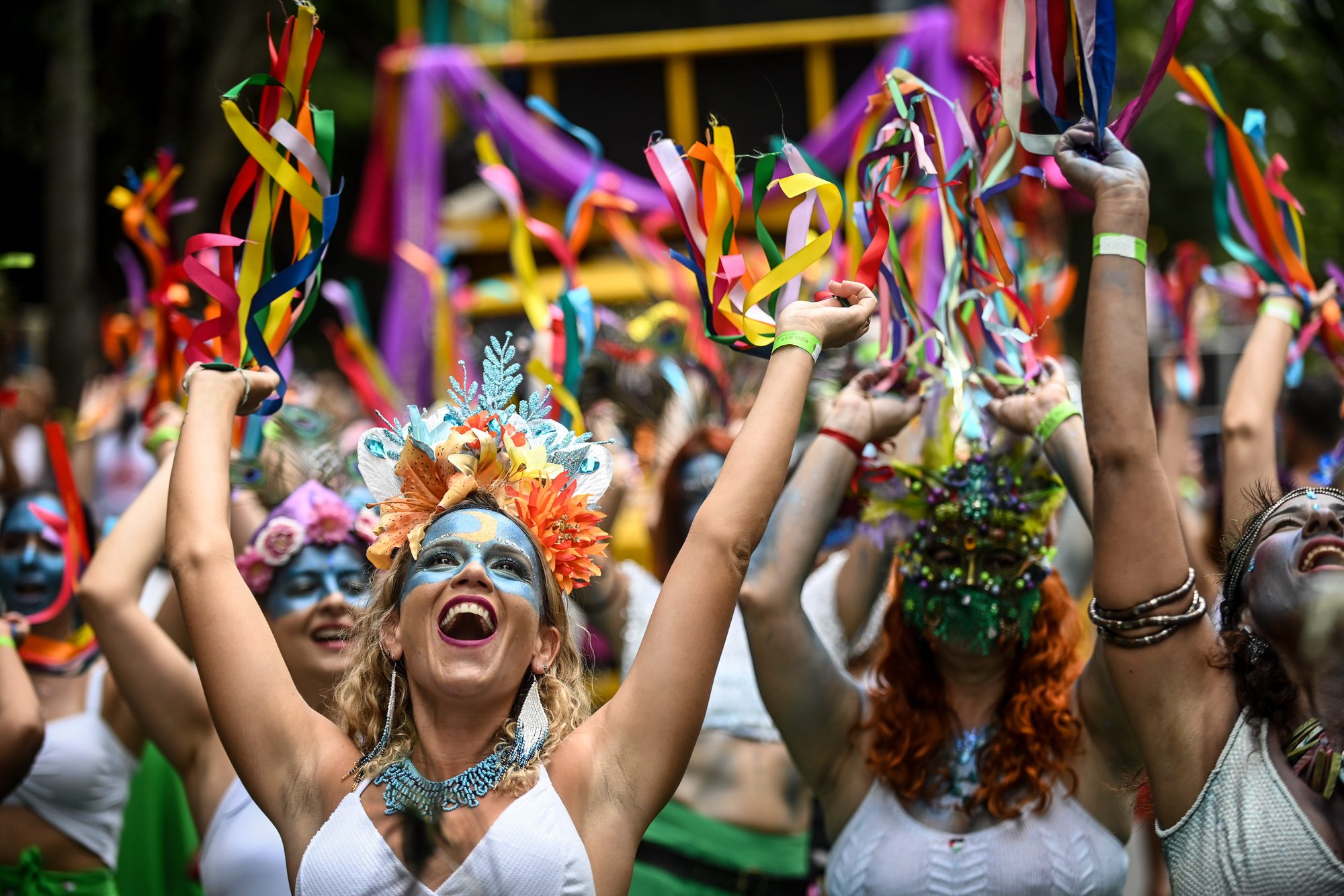 Foliãs com maquiagem azul, coroa de flores e fitas coloridas nas mãos, cantando de braços erguidos no desfile do bloco Pena de Pavão de Krishna, no bairro União, em Belo Horizonte.
