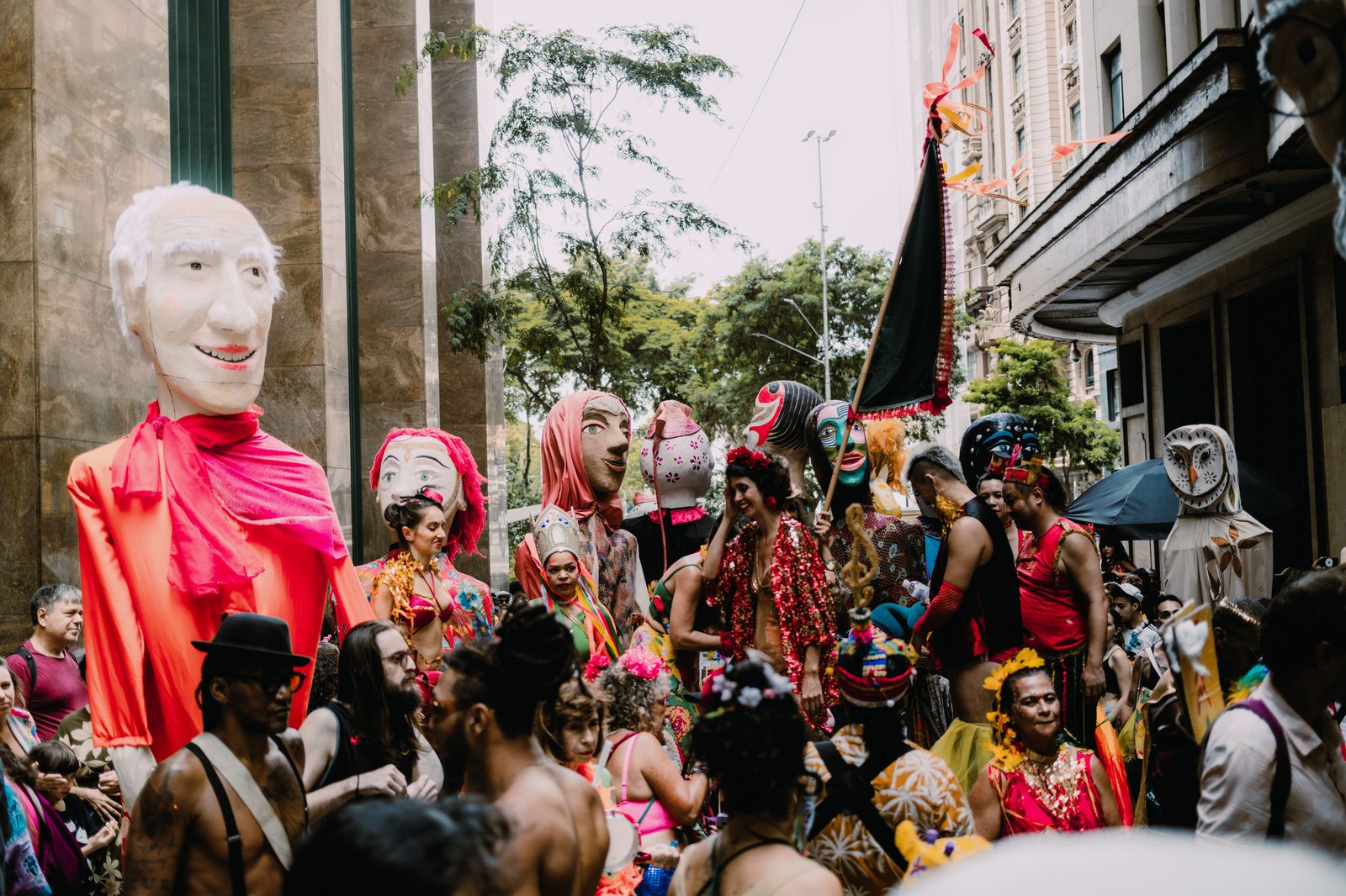 Bonecos gigantes de bloco de carnaval de rua, com rosto pintado e roupas coloridas, cercados por foliões fantasiados numa rua cercada de prédios altos.