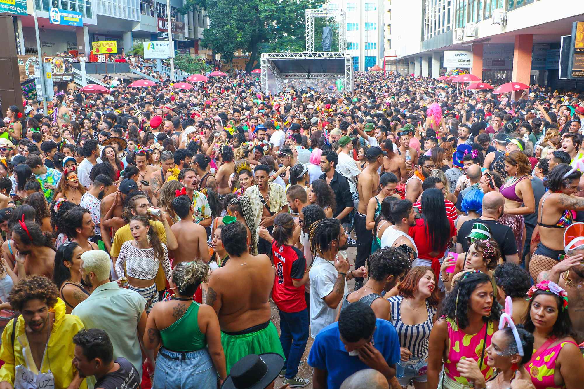 Multidão fantasiada, com tiaras, perucas coloridas e glitter, ocupando a rua entre os prédios do Setor Comercial Sul, em Brasília, com um palco de treliça metálica ao fundo e guarda-sóis vermelhos nas calçadas.