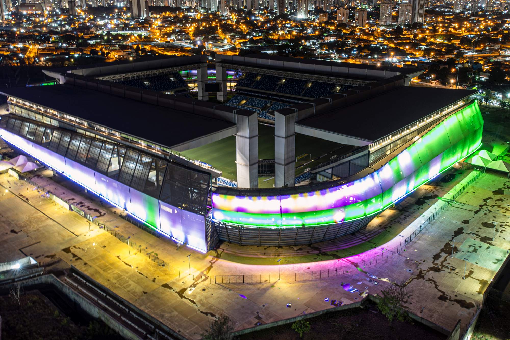 Arena Pantanal iluminada de verde, roxo e amarelo numa noite em Cuiabá, vista do alto, com as luzes da cidade ao fundo.