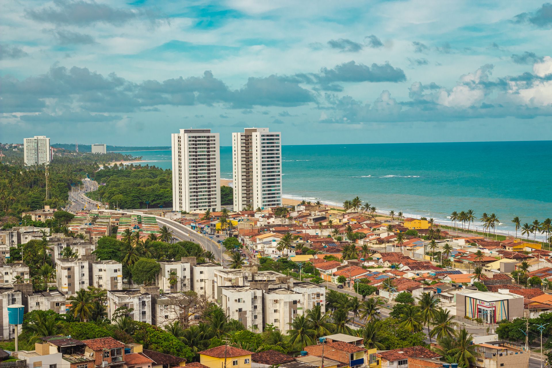 Vista da orla de Maceió durante o dia, com prédios entre o verde e o mar azul-turquesa.