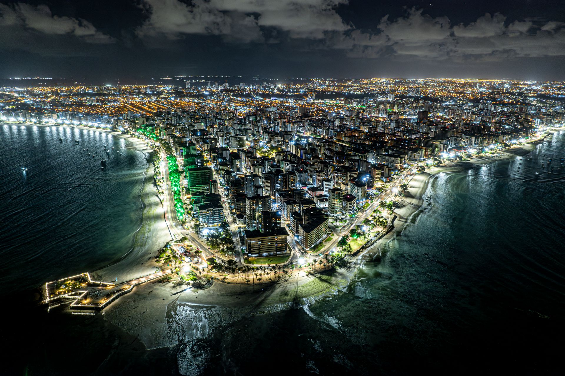 Vista aérea noturna da orla de Maceió, com as luzes dos prédios refletidas na água escura do mar.