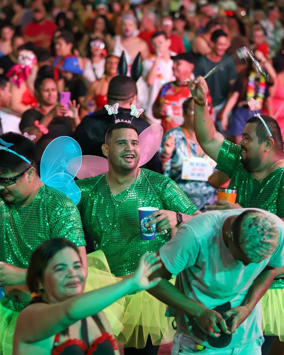Foliões dançando à noite num bloco de bairro em Manaus, com camisas verdes brilhantes, asas de tule e copos na mão, e a multidão iluminada ao fundo.