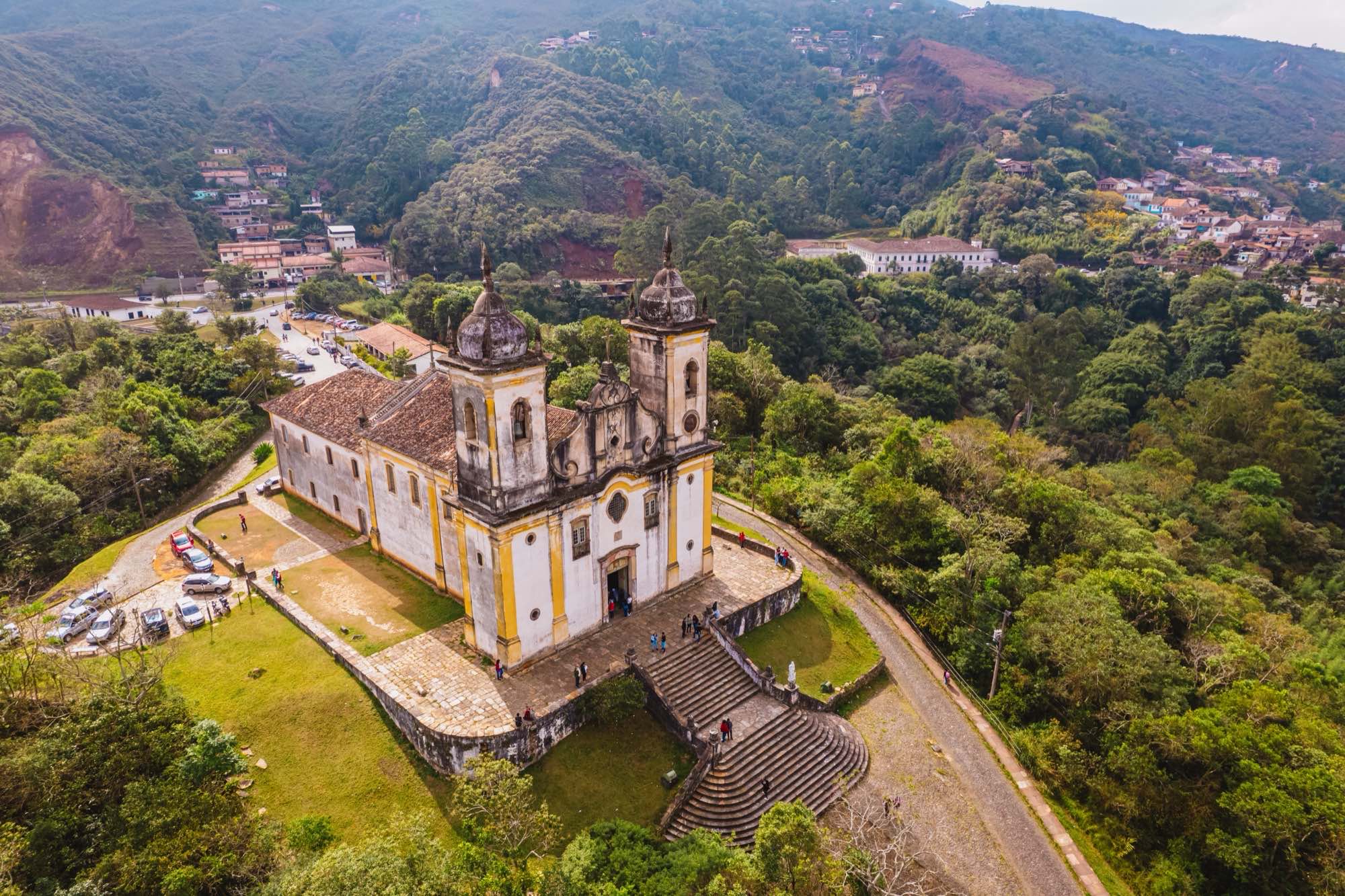 Vista aérea de uma igreja barroca branca com torres, no alto de um morro verde, em Ouro Preto, com a cidade e as montanhas ao fundo.