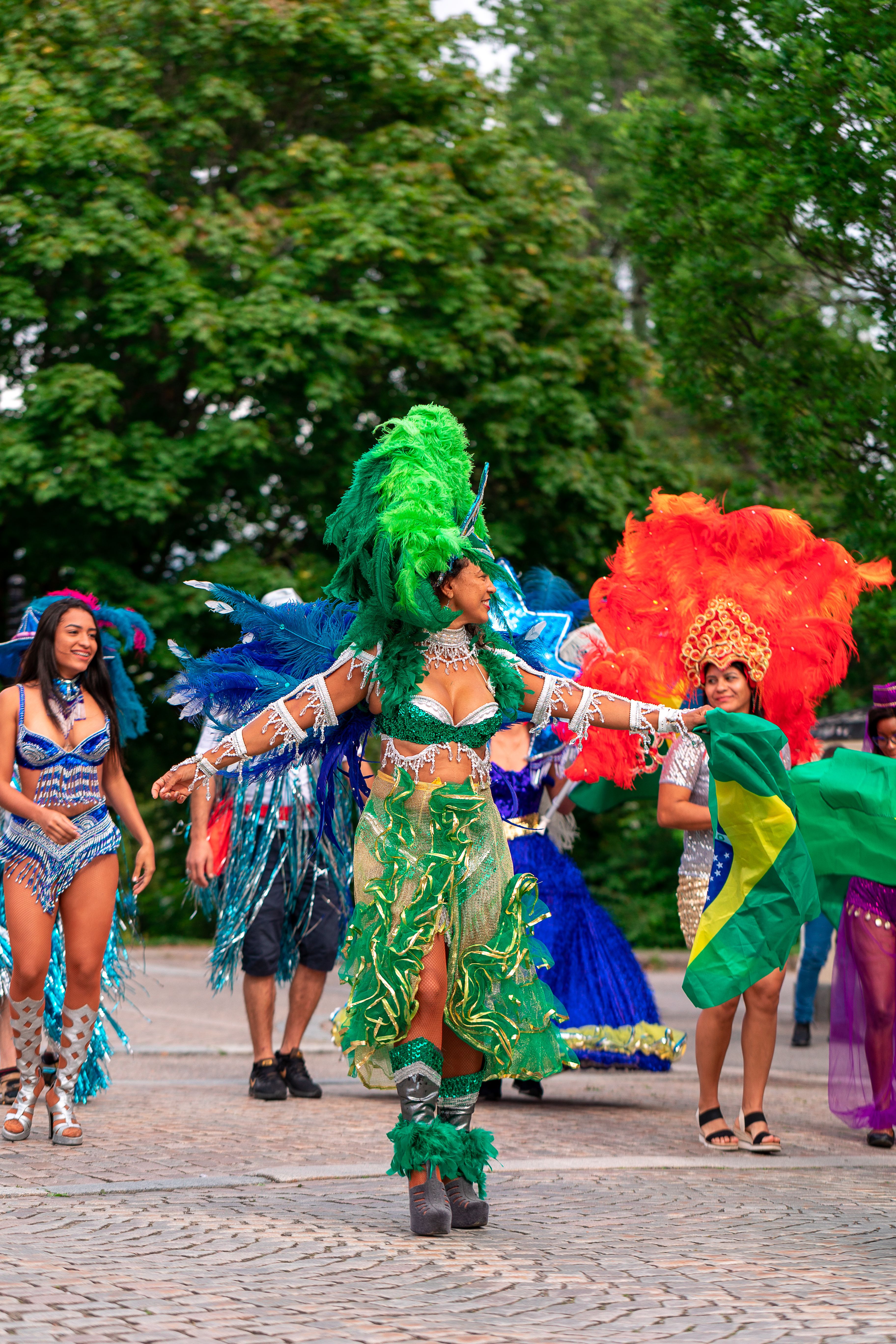 Dançarinas de carnaval com fantasia de penas verdes e laranja dançando de braços abertos numa rua de paralelepípedo, com bandeira verde e amarela ao fundo e árvores altas ladeando a via.