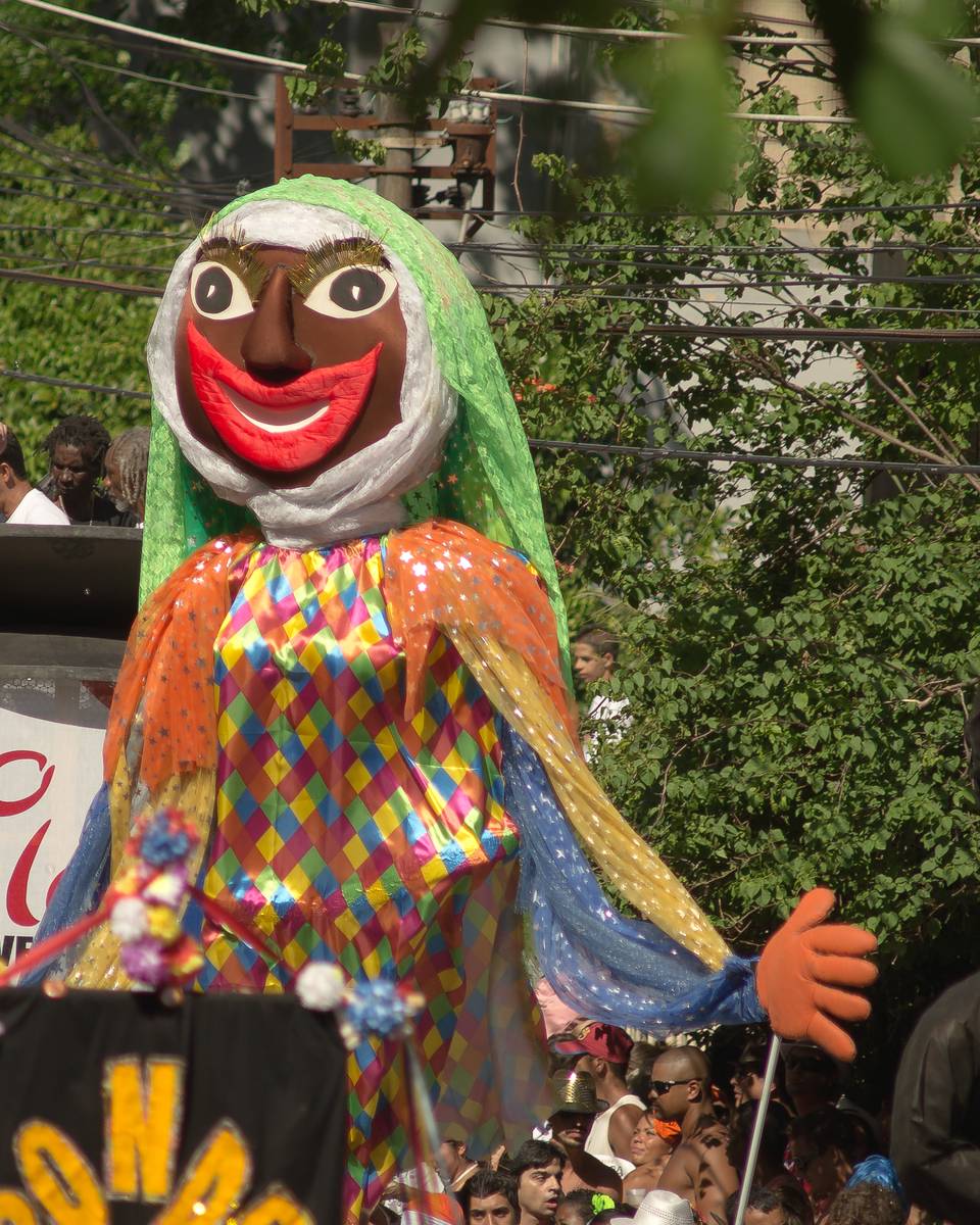 Boneco gigante de freira, de roupa colorida em losangos, acima da multidão do Bloco das Carmelitas numa rua arborizada de Santa Teresa, no Rio de Janeiro.