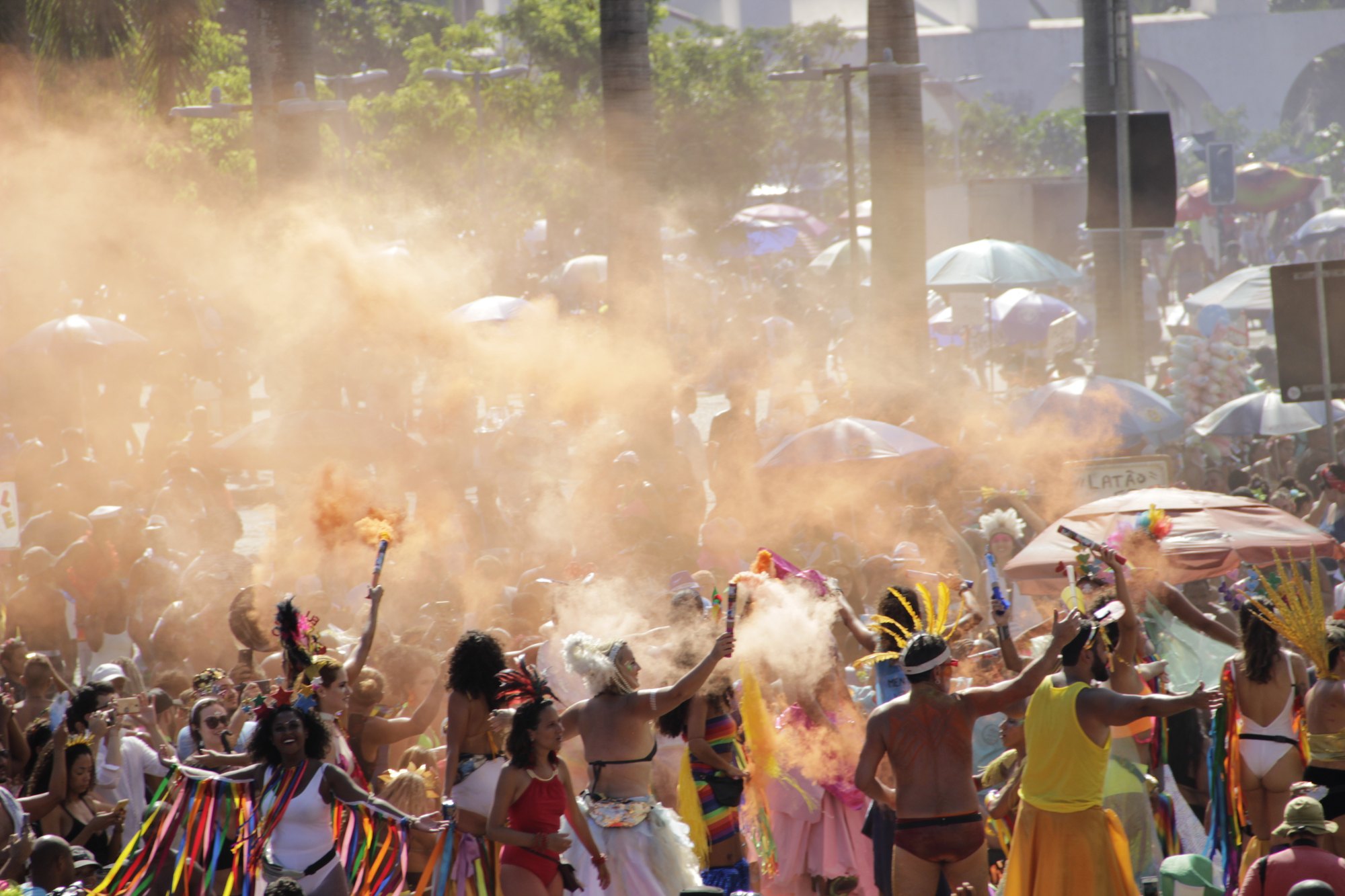Foliões fantasiados, com fitas coloridas, cocares e plumas, dançando entre fumaça laranja e guarda-sóis num bloco de Carnaval de rua no Rio de Janeiro.