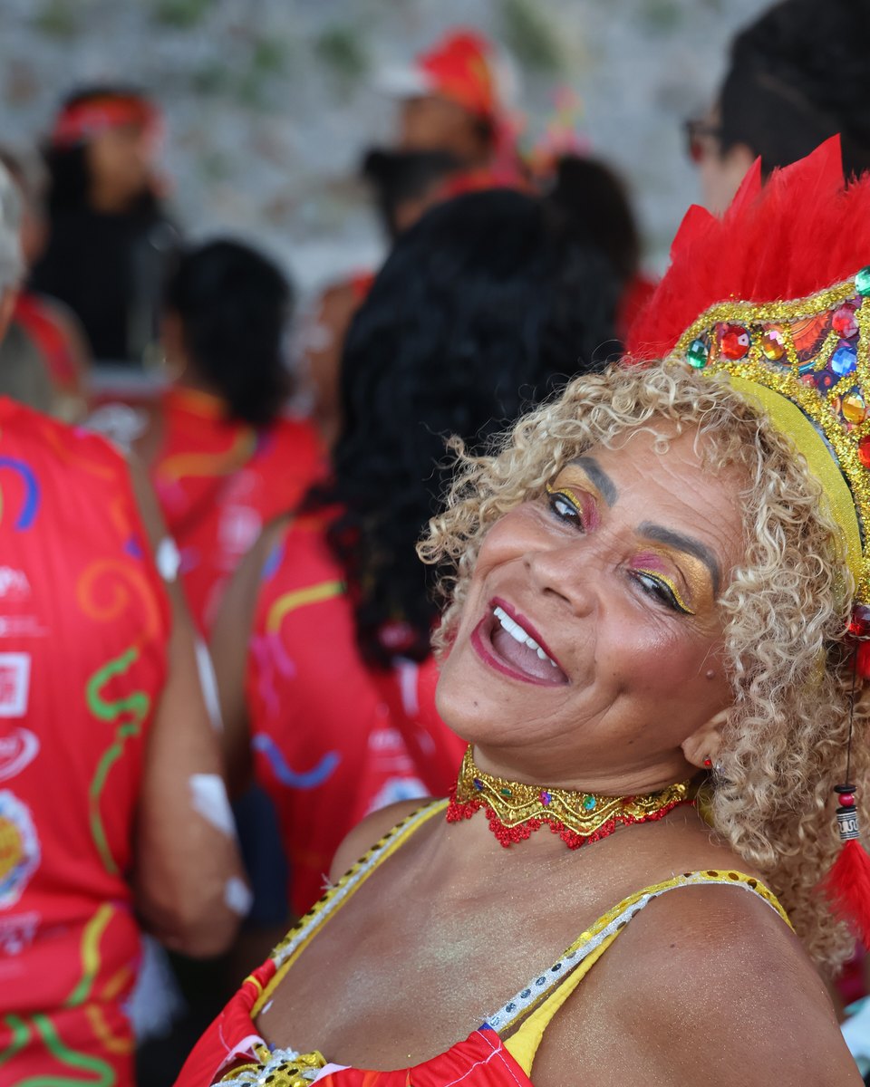 Foliã de cabelo cacheado loiro, coroa de plumas vermelhas e pedrarias, sombra dourada e gargantilha vermelha sorrindo para a câmera no Sudesb Folia, bloco de abertura do Carnaval no Santo Antônio Além do Carmo, em Salvador, com outros foliões de camiseta vermelha ao fundo.