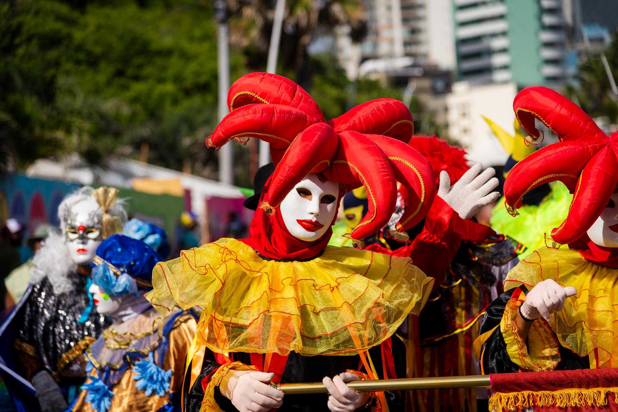 Foliões mascarados com fantasia de pierrô vermelha e amarela, gola de tule e chapéu de pontas, desfilando no Fuzuê, pré-Carnaval no circuito Barra-Ondina, em Salvador, com os prédios da orla ao fundo.