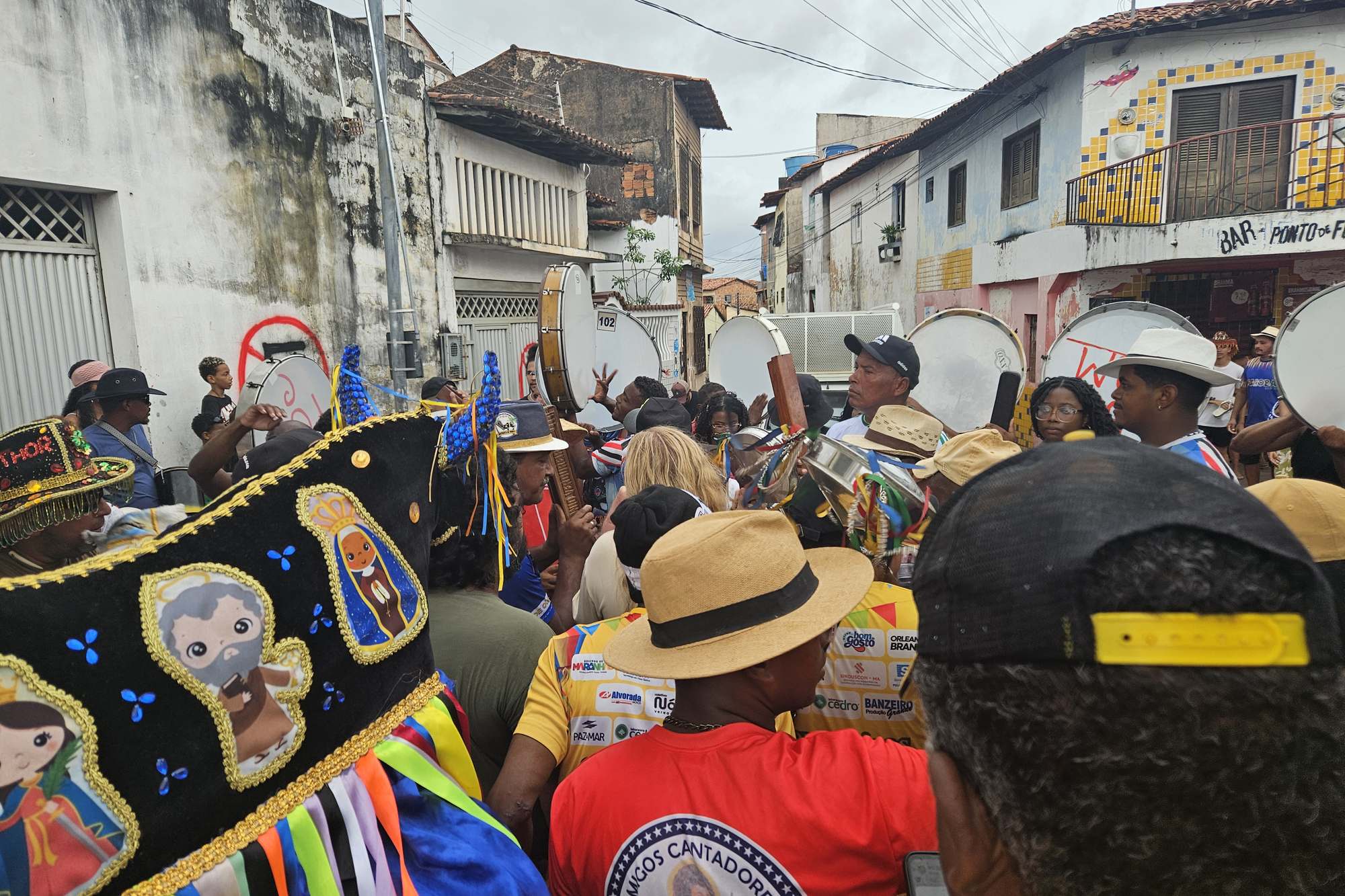 Cortejo de rua no bairro da Madre Deus, em São Luís, com brincantes de chapéu de palha, pandeirões erguidos e um boi bordado com fitas coloridas em primeiro plano, entre casarões antigos e uma fachada de azulejo amarelo.