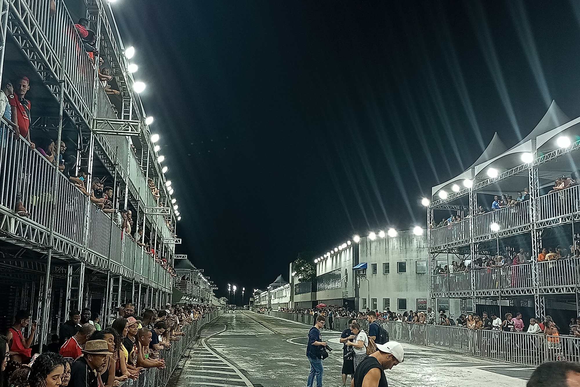 Arquibancadas de estrutura metálica cheias de público dos dois lados da pista do Sambão do Povo, em Vitória, iluminadas à noite antes de um desfile.