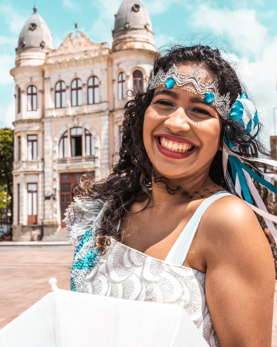 Passista de frevo sorrindo, com tiara de pedrarias, fitas azuis no cabelo e roupa branca bordada, no calçadão do Marco Zero, no Bairro do Recife, com o prédio histórico de cúpulas e outros casarões ao fundo.