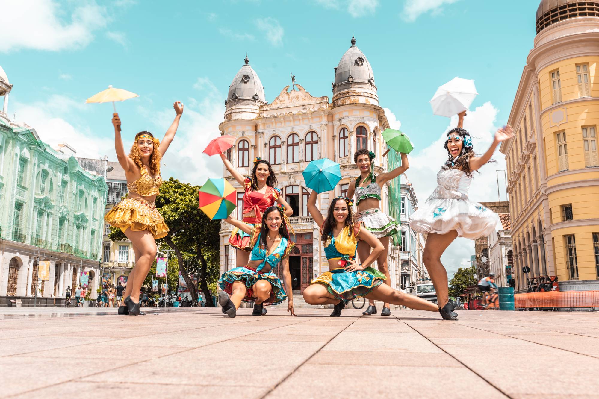 Seis passistas de frevo com saias de babado coloridas e sombrinhas de frevo erguidas, posando no calçadão do Marco Zero, no Bairro do Recife, com prédios históricos amarelo, verde e de cúpulas ao fundo.