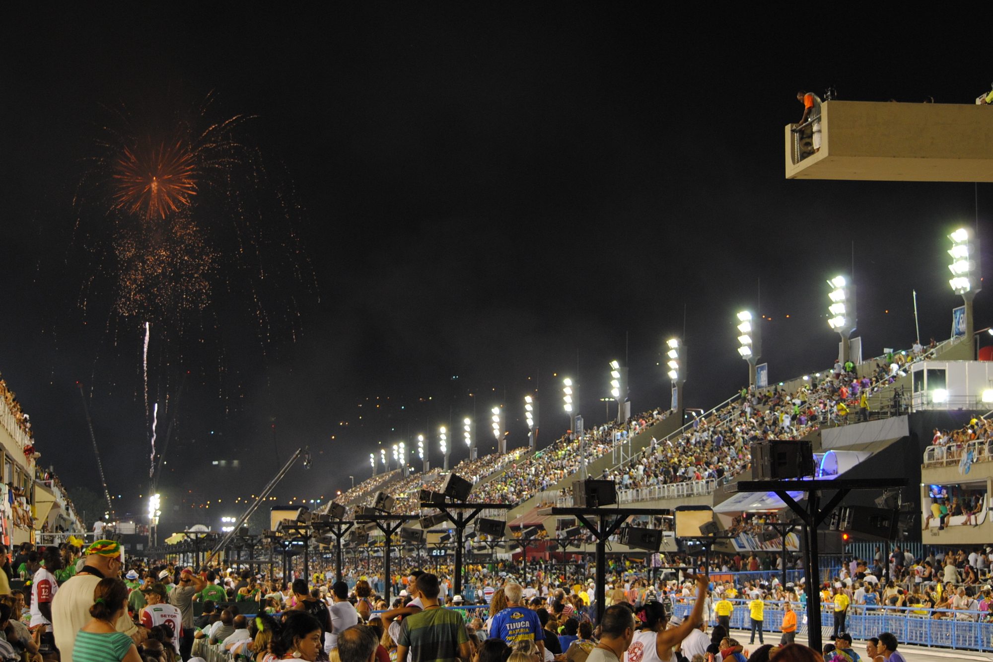 Público na pista e nas arquibancadas iluminadas do Sambódromo da Marquês de Sapucaí, no Rio de Janeiro, à noite, com fogos de artifício no céu.