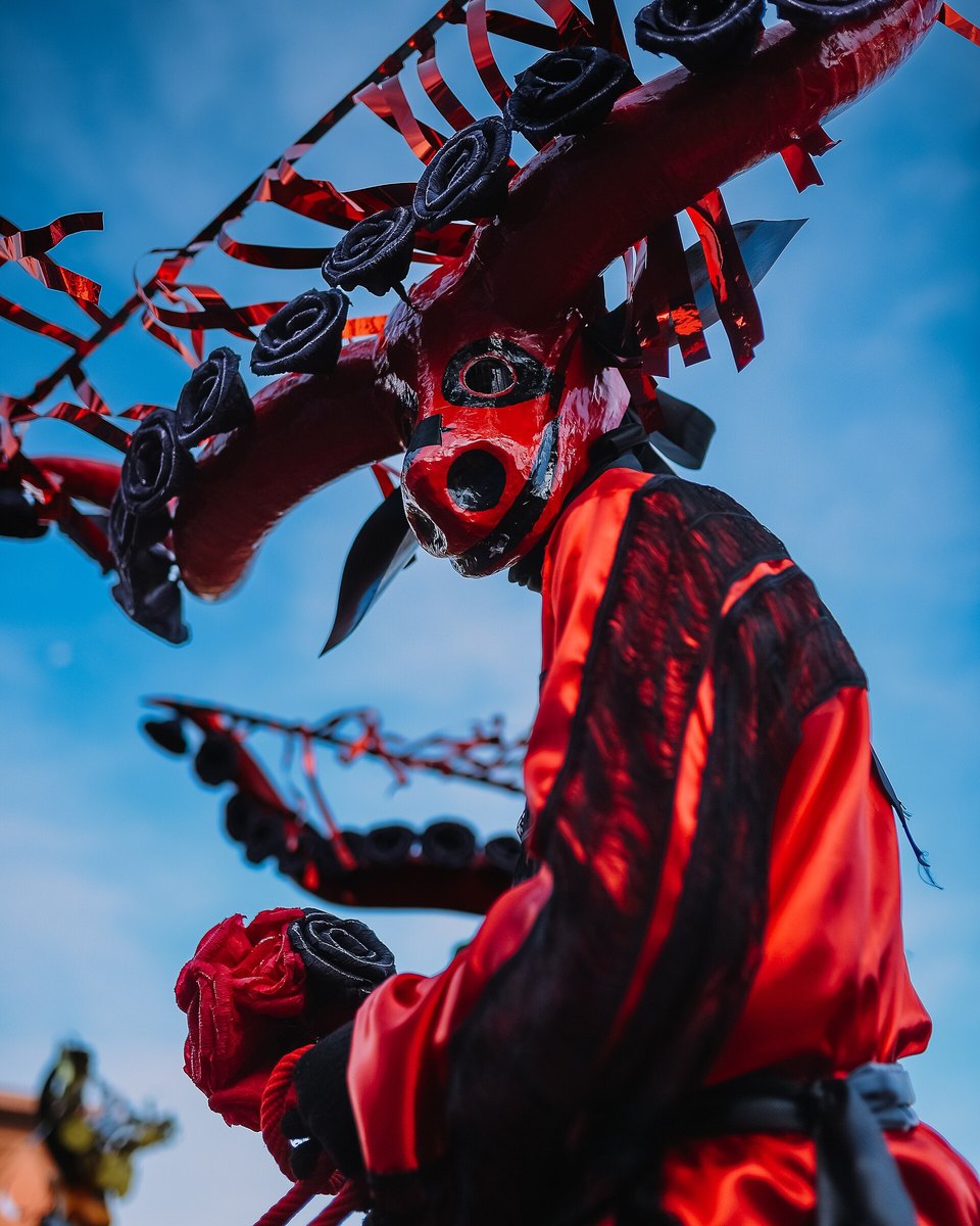 Mascarado das Cavalhadas de Pirenópolis com máscara vermelha, roupa de cetim vermelho e preto e um grande enfeite de rosas pretas e fitas vermelhas na cabeça, contra o céu azul.