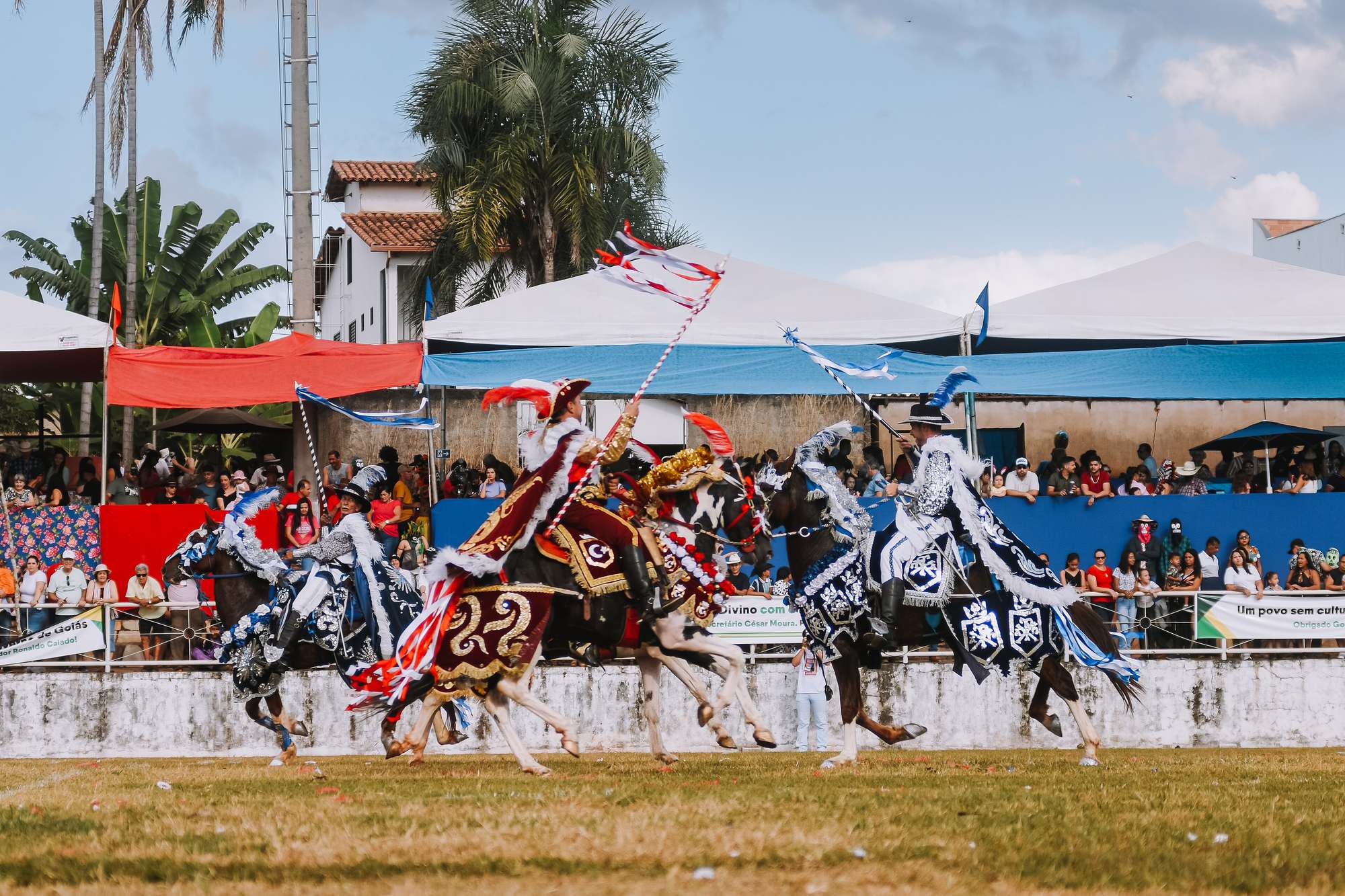 Cavaleiros das Cavalhadas de Pirenópolis, com roupas e mantas de cavalo bordadas em azul, branco, vermelho e dourado, galopando com lanças enfeitadas de fitas diante da arquibancada cheia de público, sob tendas brancas e palmeiras.