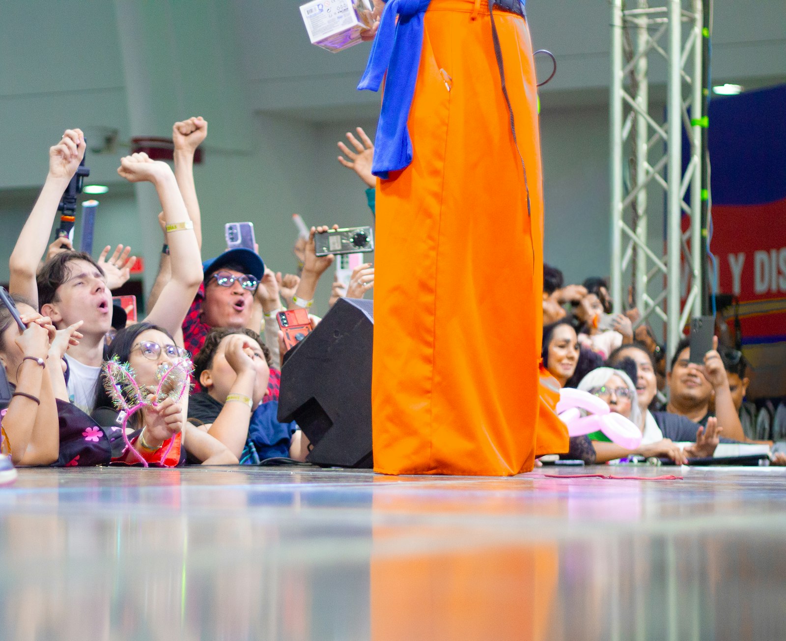 Fãs na grade de um palco de convenção de cultura pop gritando, com os braços erguidos, e filmando com o celular um cosplayer de roupa laranja.