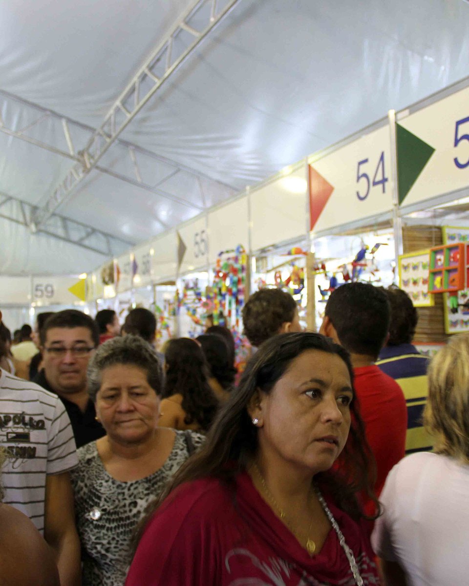 Corredor lotado da feira de artesanato de miriti em Belém, com visitantes caminhando entre estandes numerados e brinquedos de pássaros e aviões coloridos pendurados.