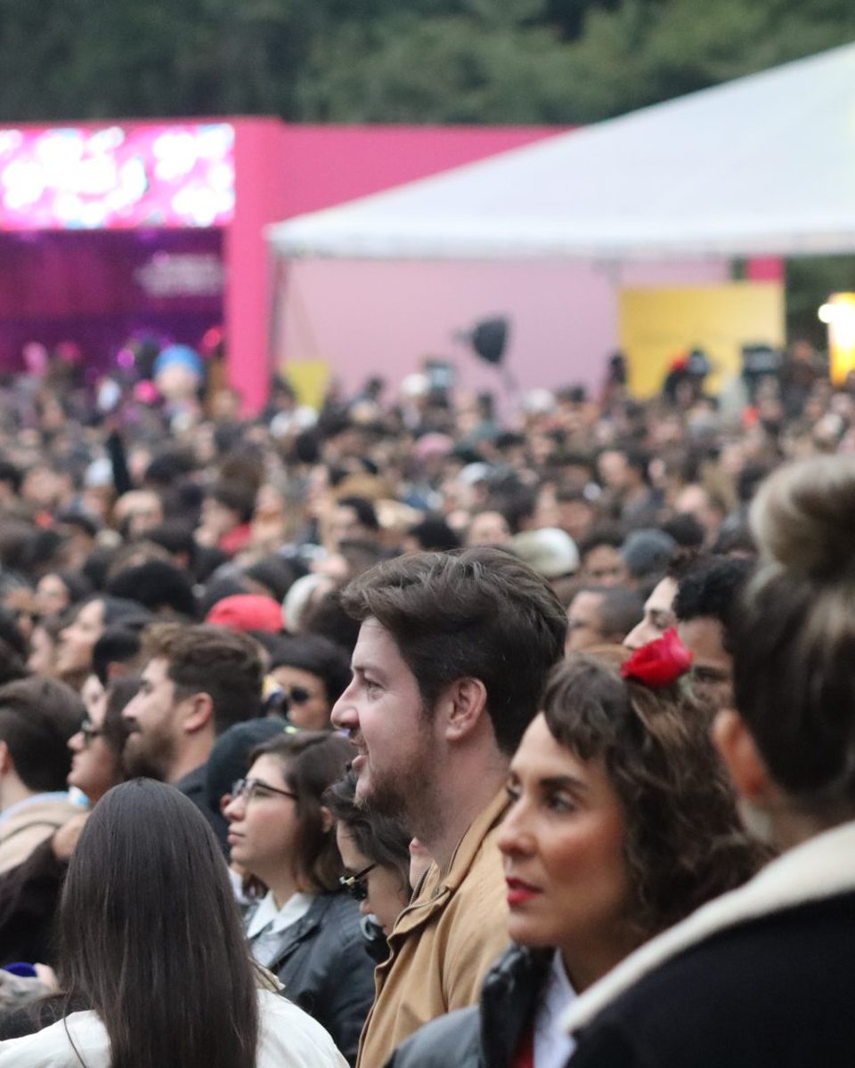 Rostos do público do Coolritiba 2026 na Pedreira Paulo Leminski, em Curitiba: rapaz de jaqueta caramelo, mulher de batom vermelho com uma flor vermelha no cabelo e casaco de gola de pelúcia, e a multidão diante de uma tenda rosa ao fundo.