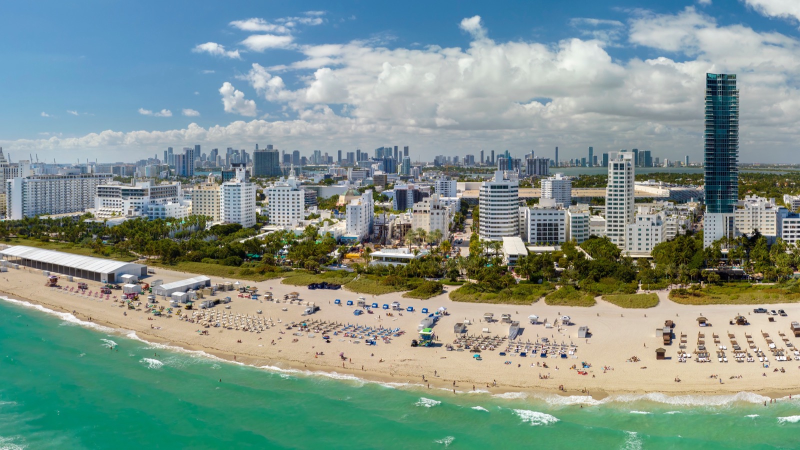 Vista aérea de South Beach, em Miami Beach, com banhistas e guarda-sóis na areia, o mar verde-água e o skyline de Miami ao fundo.