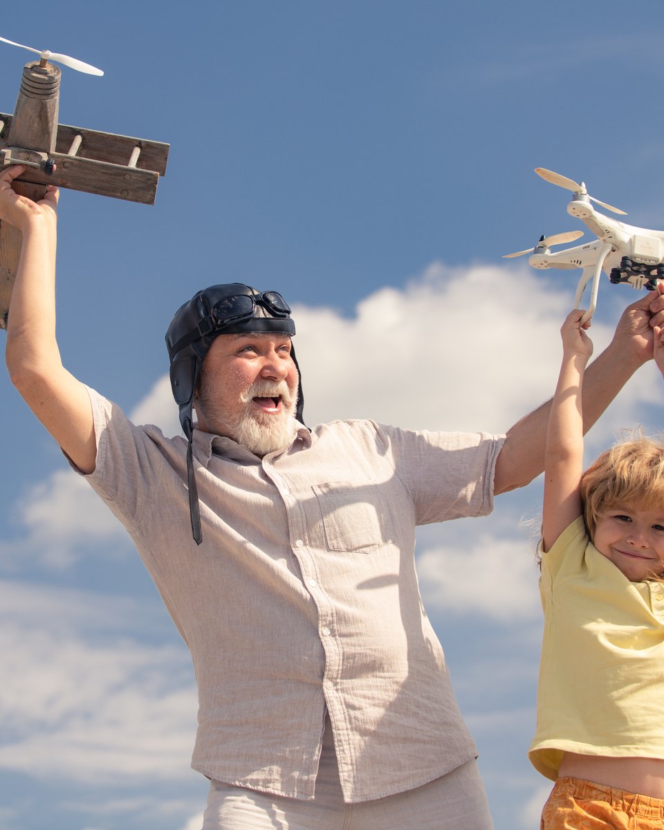 Avô de barba branca e capacete de aviador ergue um avião de madeira ao lado do neto, que segura um drone no alto, contra o céu azul.