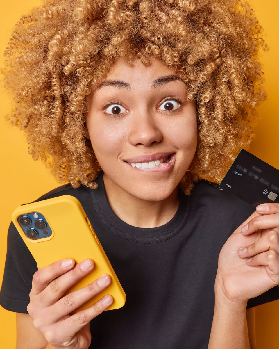 Mulher de cabelo cacheado e camiseta preta, com expressão de surpresa, segurando o celular e um cartão de crédito diante de uma parede amarela.