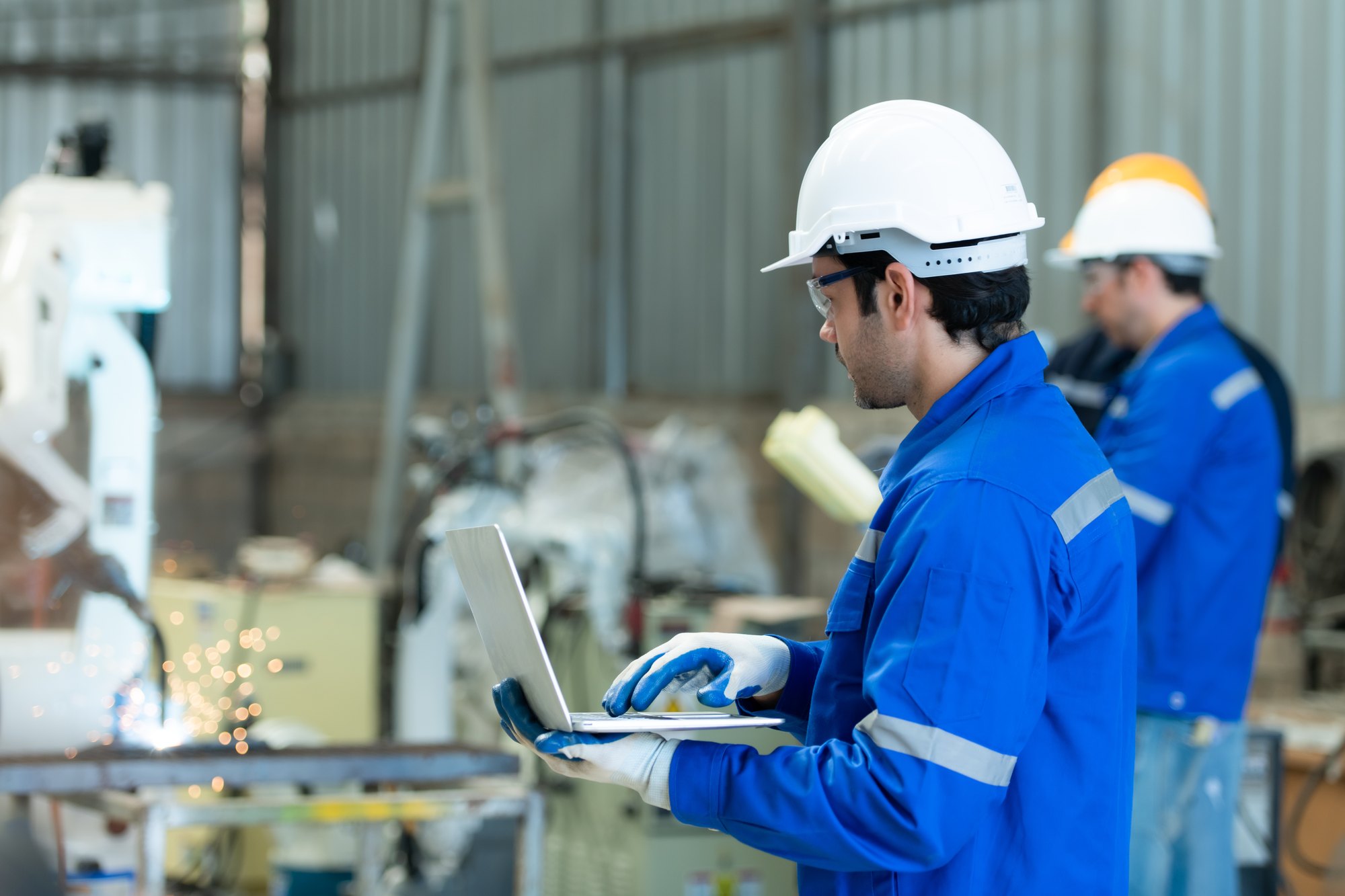Engenheiro de capacete branco e uniforme azul segurando um notebook ao lado de um robô de solda em operação, com faíscas, num galpão industrial; um colega de capacete amarelo ao fundo.