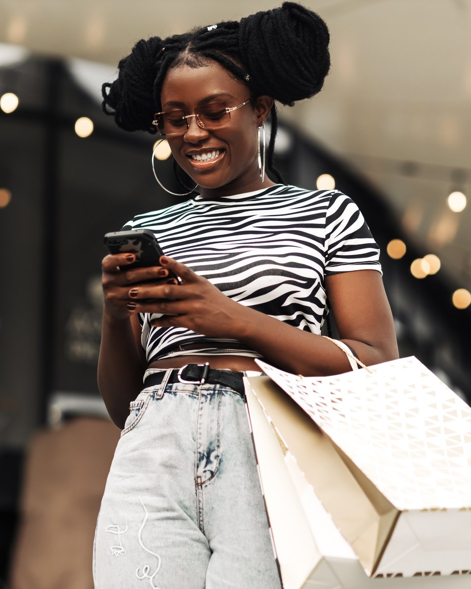 Mulher de óculos e blusa listrada em preto e branco sorrindo para o celular, com sacolas de compras no ombro, num corredor comercial com luzes penduradas.