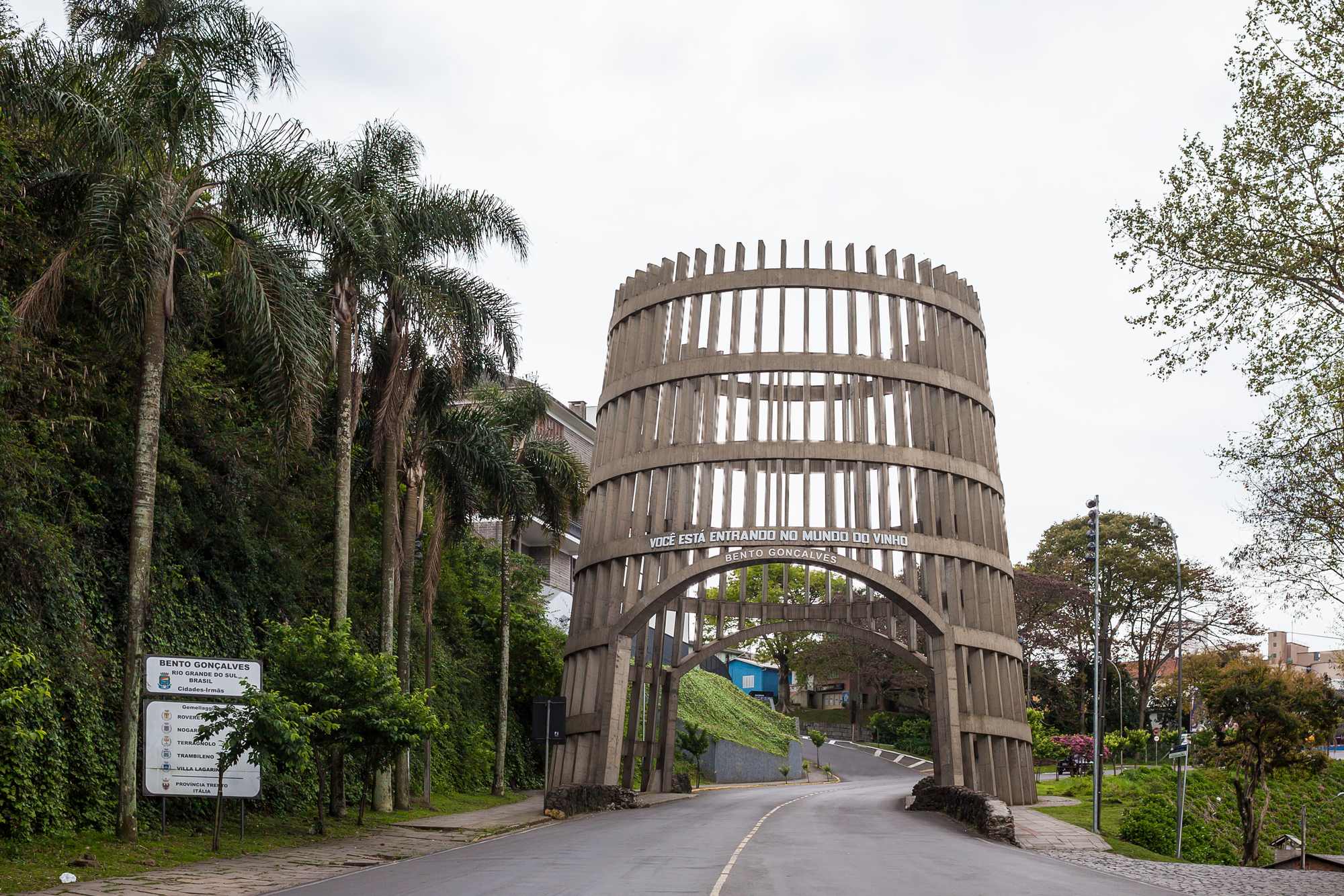 Pórtico de entrada de Bento Gonçalves em forma de pipa de vinho, com a inscrição "Você está entrando no mundo do vinho", sobre a avenida arborizada com palmeiras, num dia nublado.