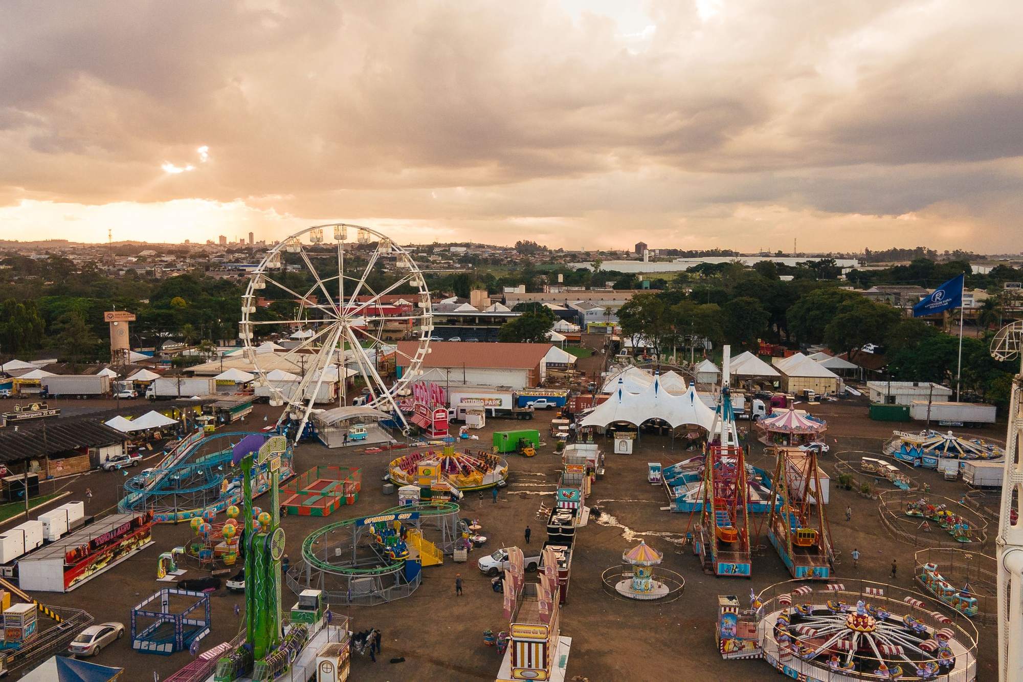 Vista aérea do parque de diversões da ExpoLondrina ao entardecer, no Parque Ney Braga, em Londrina, com a roda-gigante, montanhas-russas, carrosséis e tendas brancas sob um céu dourado e a cidade ao fundo.