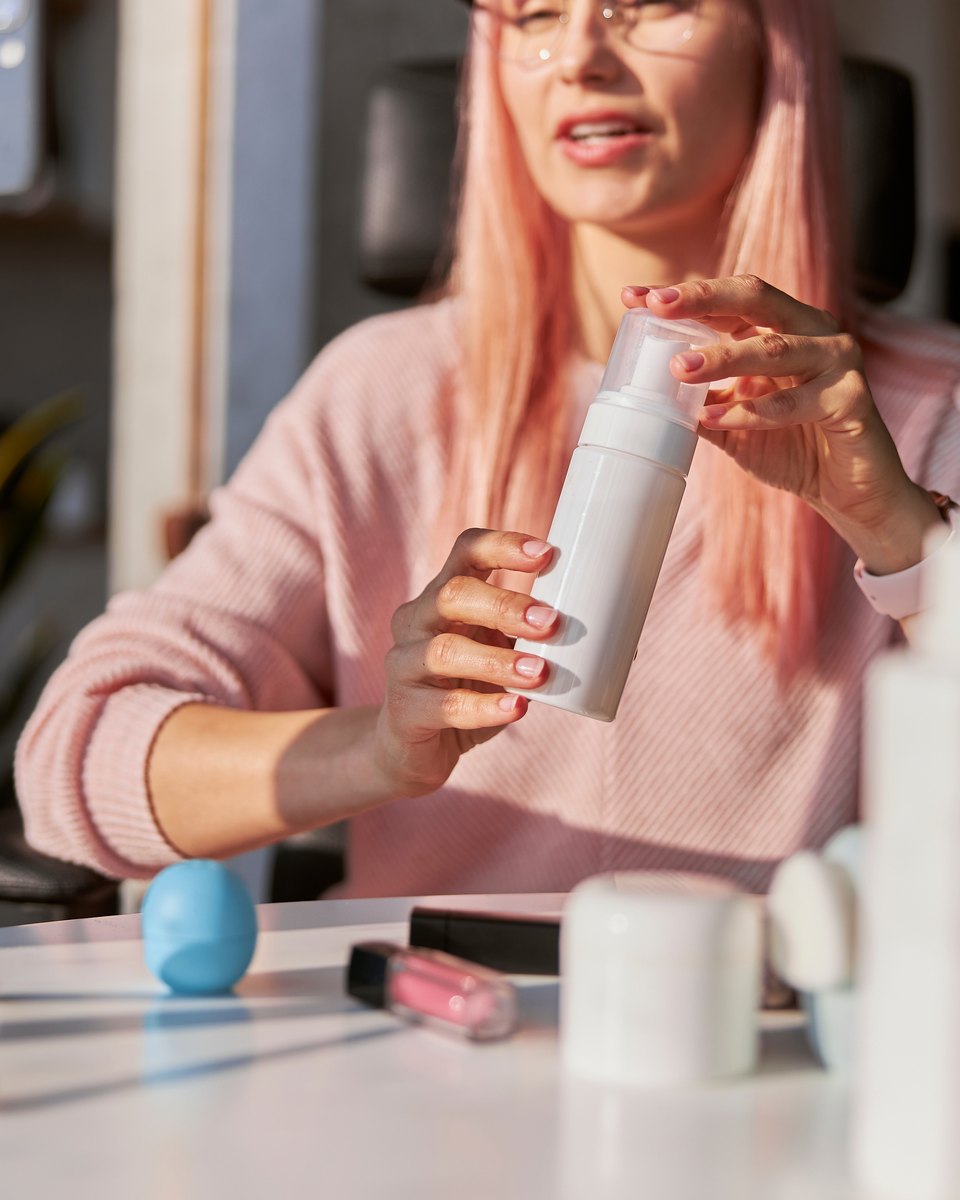 Jovem de cabelo rosa e blusa rosa mostrando um frasco branco sem rótulo para o celular preso num tripé, numa mesa com potes e bisnagas de cosméticos.
