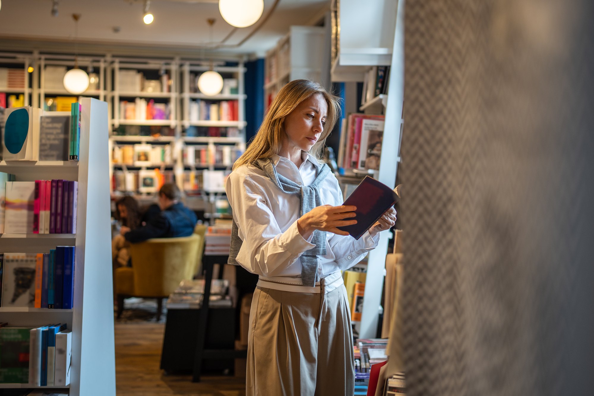 Mulher de camisa branca e suéter cinza nos ombros folheando um livro de capa vinho entre estantes de uma livraria, com leitores sentados numa poltrona amarela ao fundo.