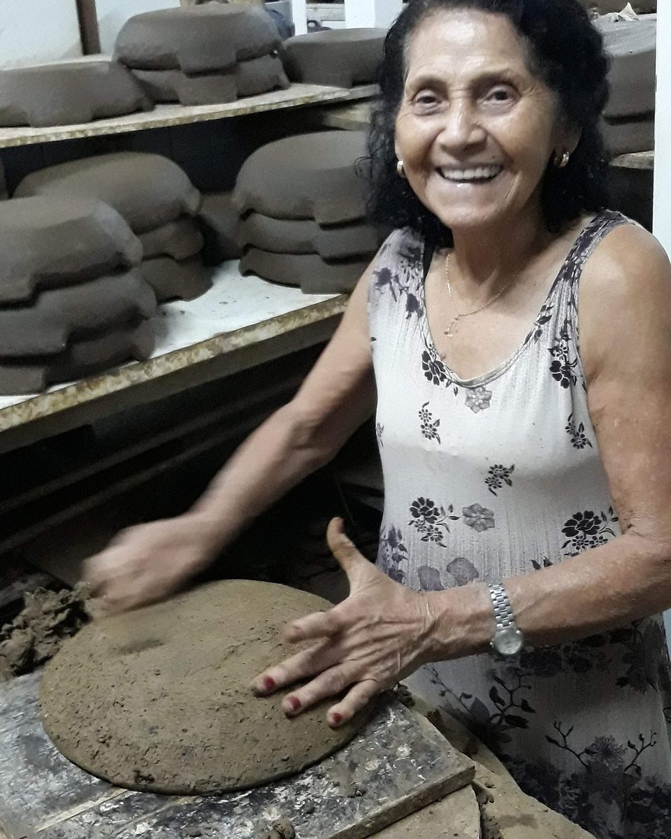 Paneleira da Associação das Paneleiras de Goiabeiras, em Vitória, sorrindo enquanto alisa com as mãos uma panela de barro sobre a bancada, com prateleiras de peças cruas secando ao fundo.
