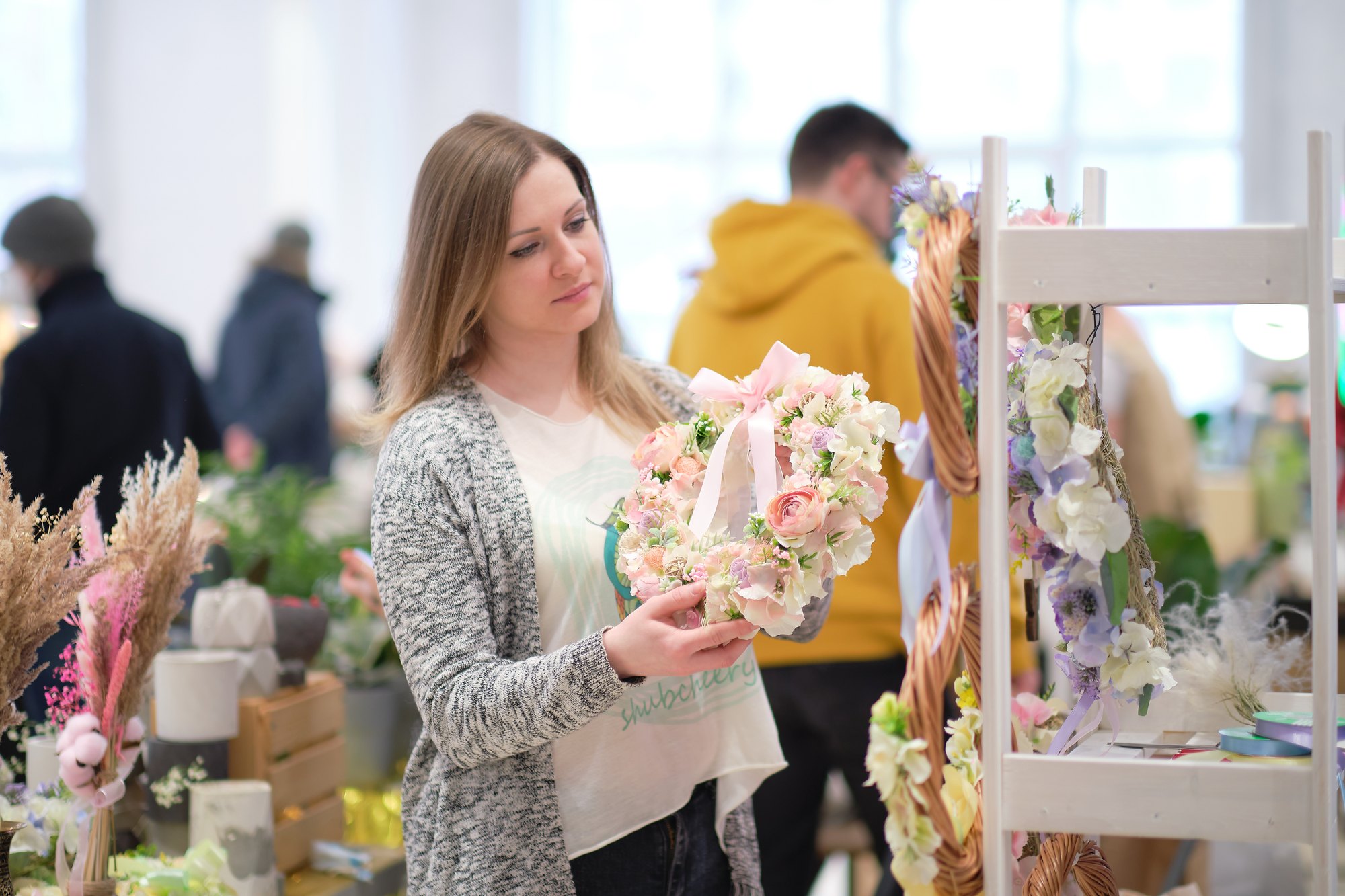 Mulher de cardigã cinza segura uma guirlanda de flores com laço rosa em uma feira de artesãos coberta, entre prateleiras de peças feitas à mão, vasos e flores secas, com outros visitantes circulando ao fundo.