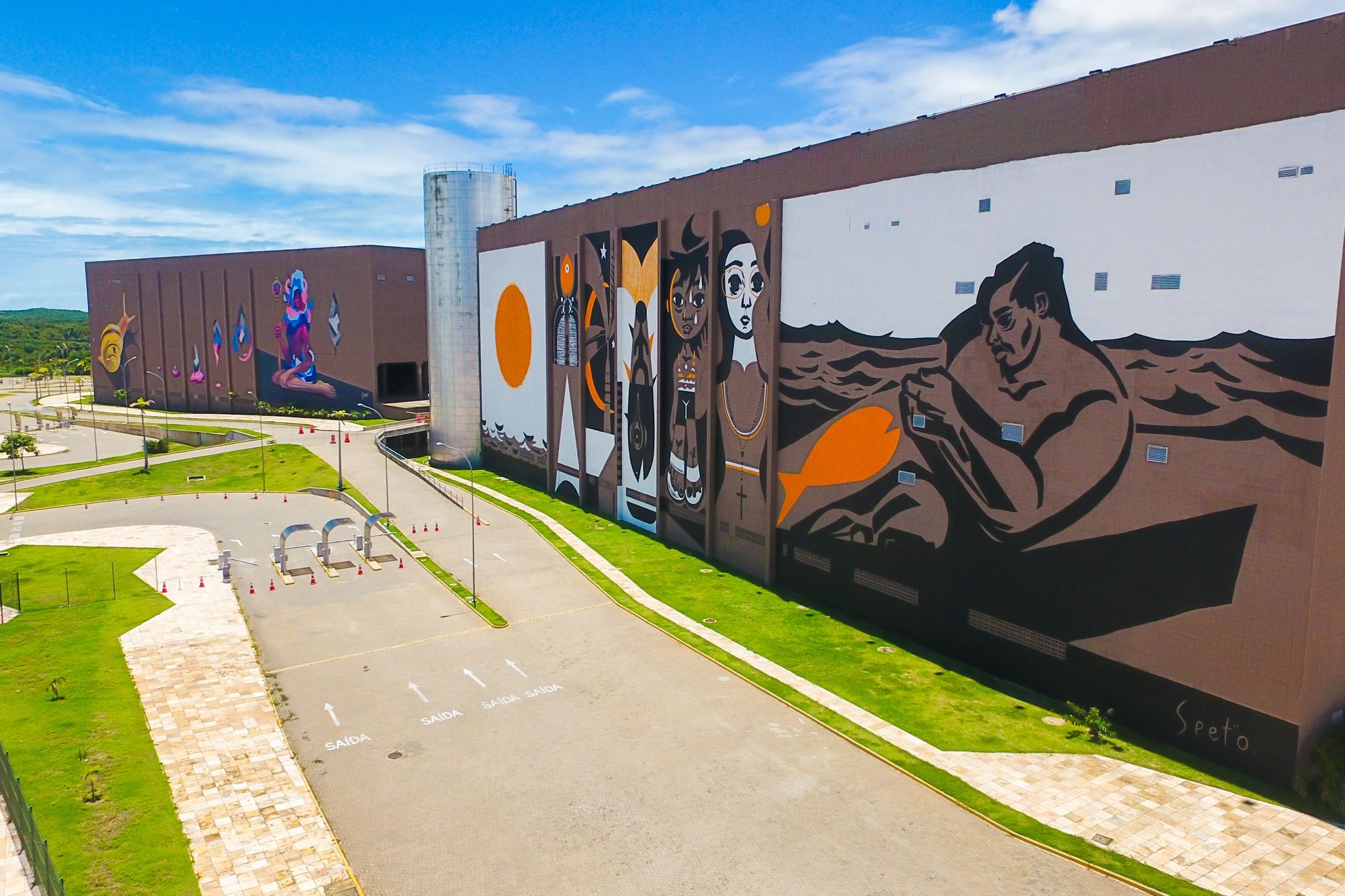 Vista de drone da fachada do Centro de Eventos do Ceará, em Fortaleza, com um grande mural colorido pintado na parede, gramado e estacionamento em frente e céu azul.