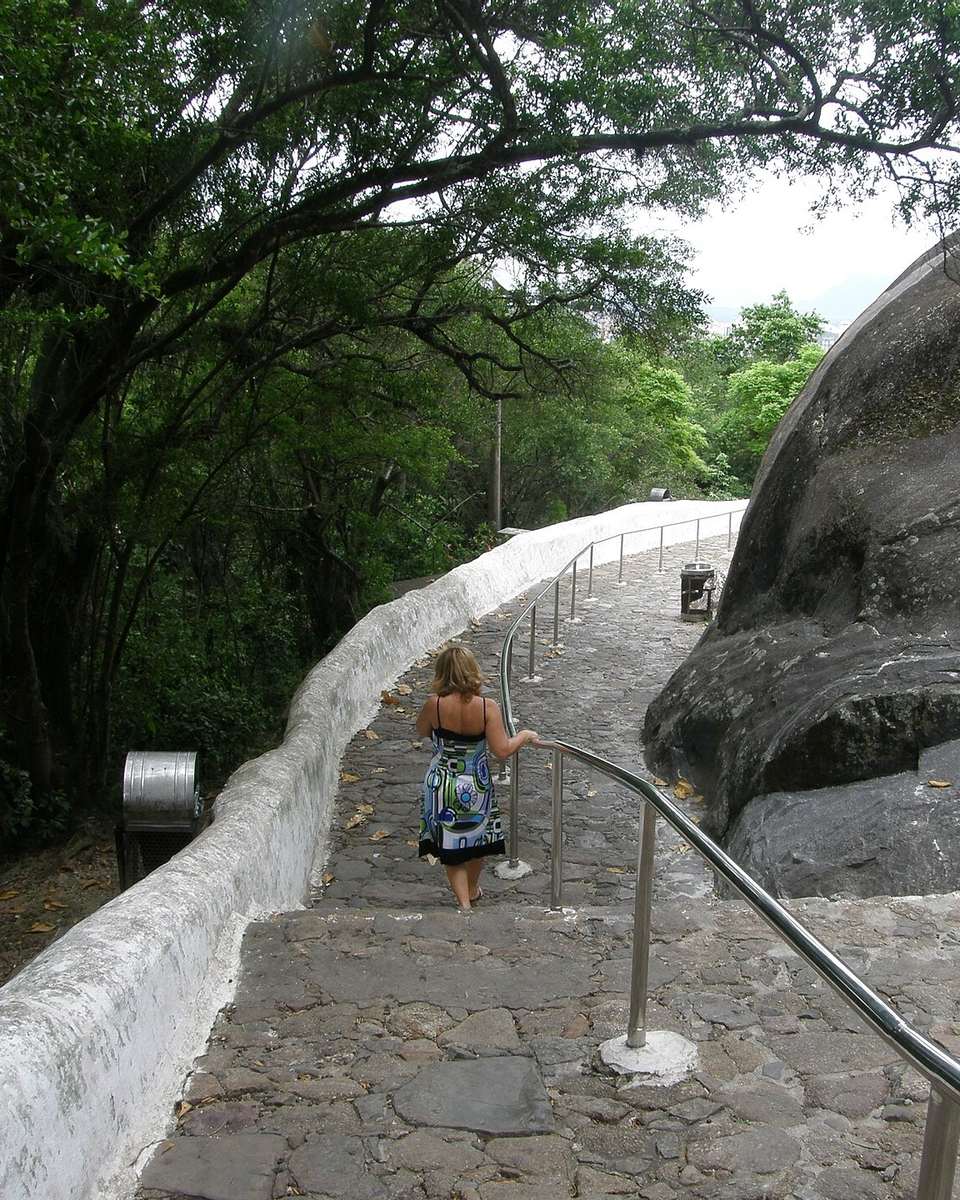 Mulher de mochila descendo sozinha a ladeira de pedras do Convento da Penha, em Vila Velha, entre o muro branco, o corrimão e o paredão de rocha.