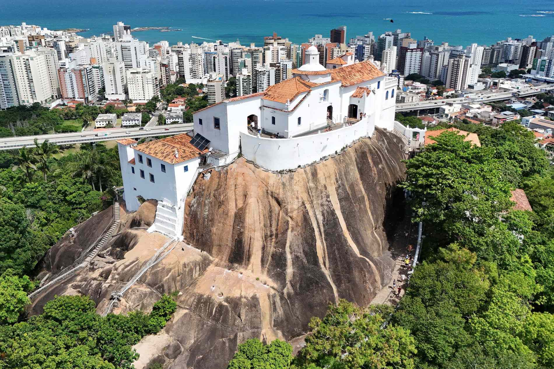 Vista aérea do Convento da Penha, conjunto branco de telhado alaranjado no alto de um rochedo cercado de mata, com os prédios de Vila Velha e o mar ao fundo.